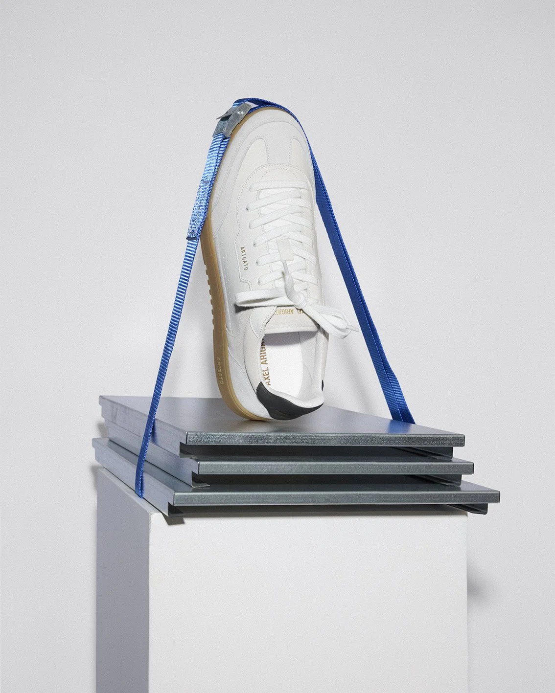 A single white sneaker with gold accents placed on a stack of gray books, secured with a blue strap, against a plain background.