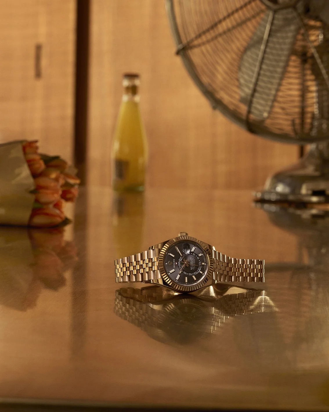 A Rolex with a black dial lying on a reflective wooden surface, with objects including a fan, a bottle, and a wrapped bouquet of flowers in the background