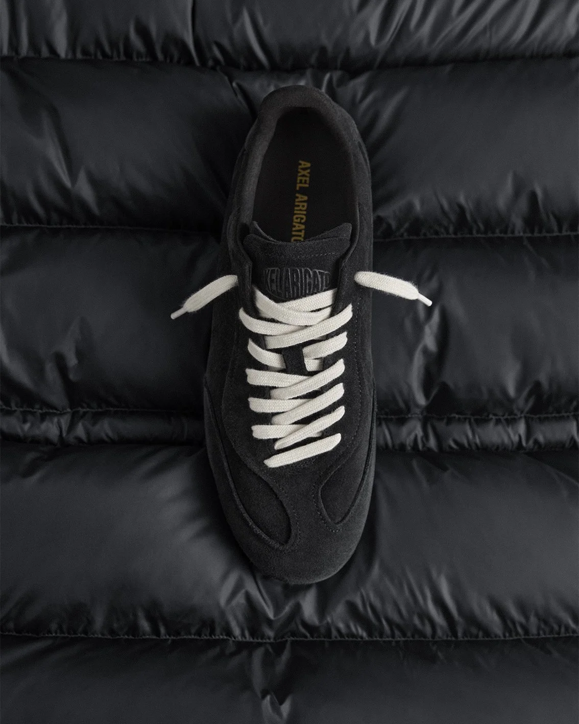 A black suede sneaker with white laces resting on a black quilted surface.