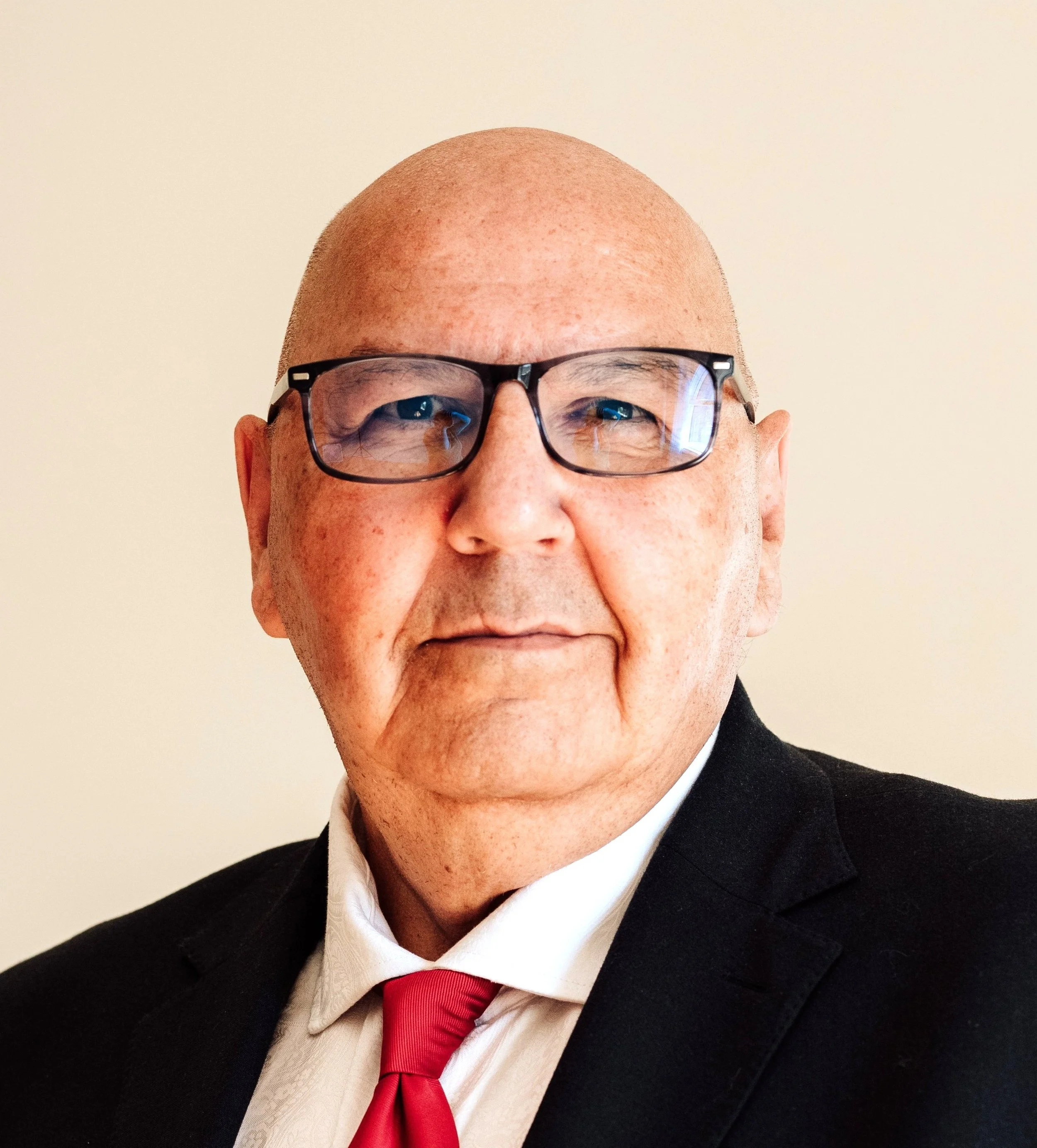 Close-up of a man's face wearing glasses, dressed in a suit with a white shirt and red tie, standing against a plain light background.