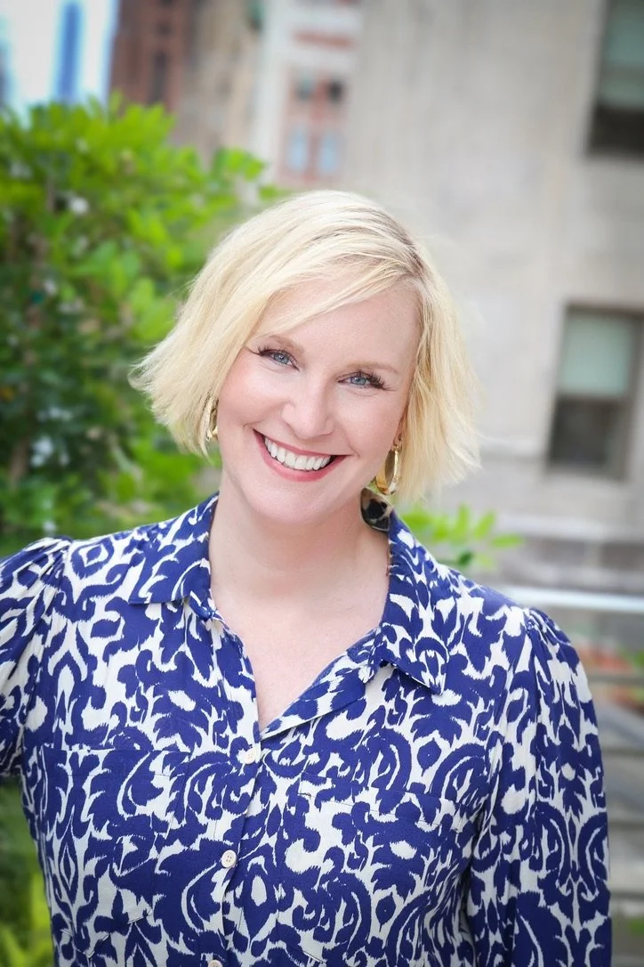 A St Gorge Wealth client with short blonde hair, wearing a blue and white patterned blouse, smiling outdoors with a green bush and building in the background.
