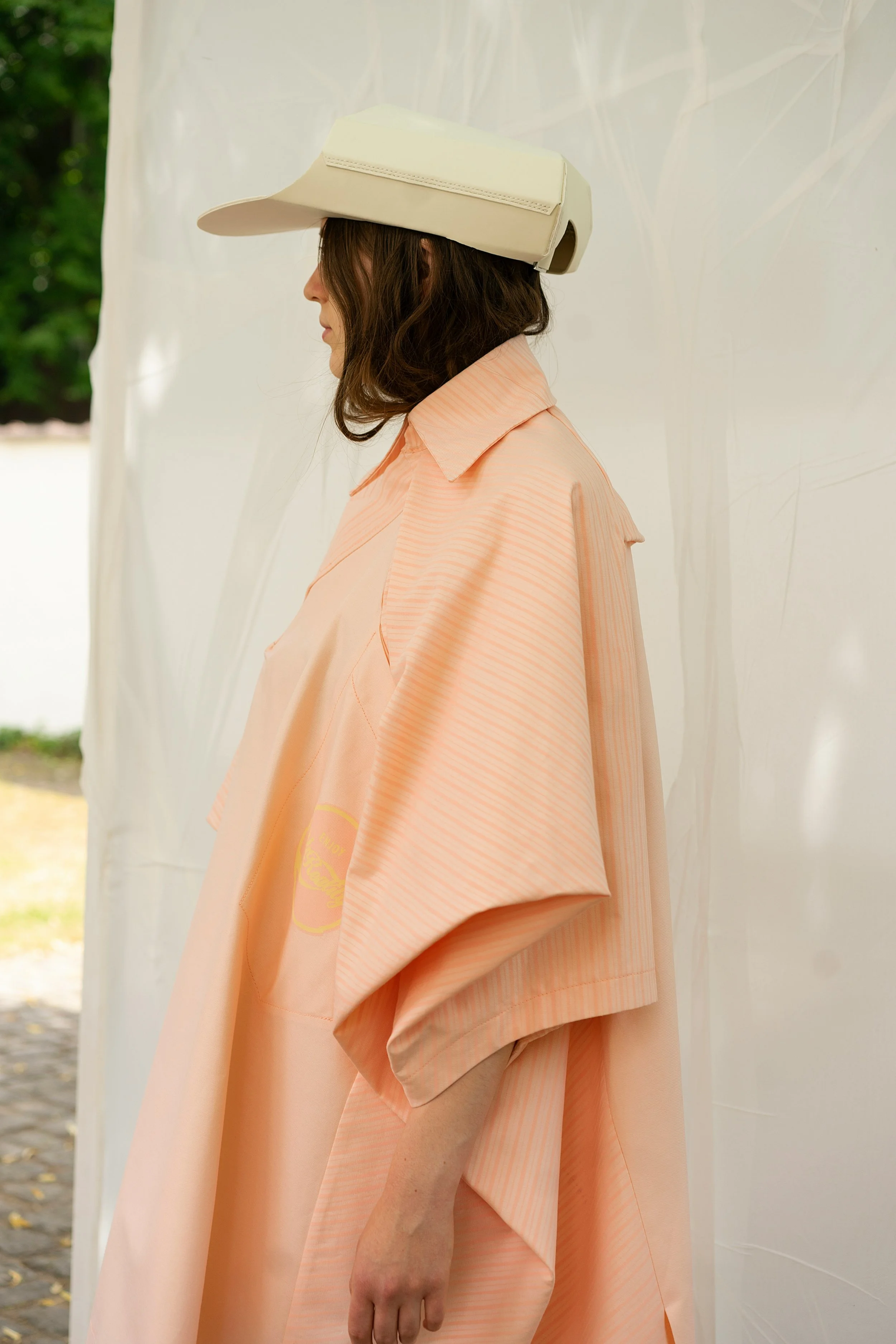 Una persona di profilo indossa un ampio abito color pesca e un cappello chiaro dal design geometrico. La luce  naturale e lo sfondo essenziale creano un'atmosfera di quiete e osservazione. L'immagine accompagna la lista d'attesa di Pattern Mirror.