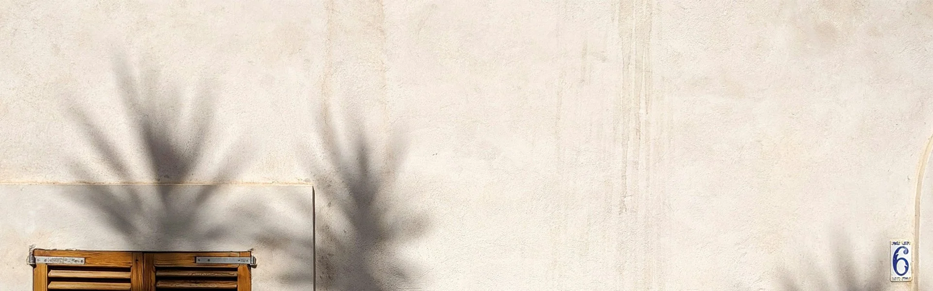 Muro di una casa con ombra di una pianta e una finestrella in legno, numero 6 in basso a destra.