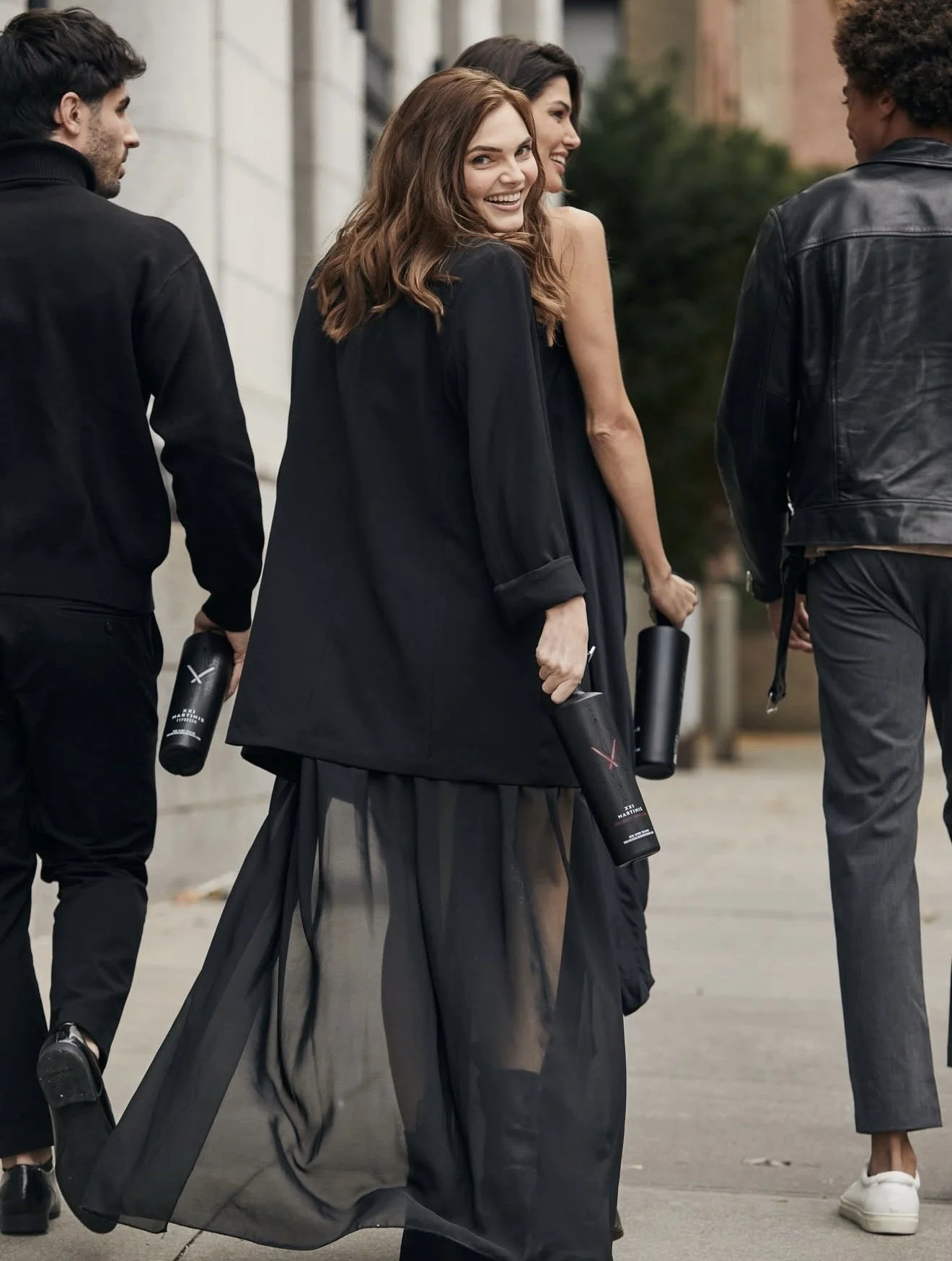 A group of four young adults walking outdoors, dressed in stylish black clothing, holding black water bottles, one woman smiling at the camera.