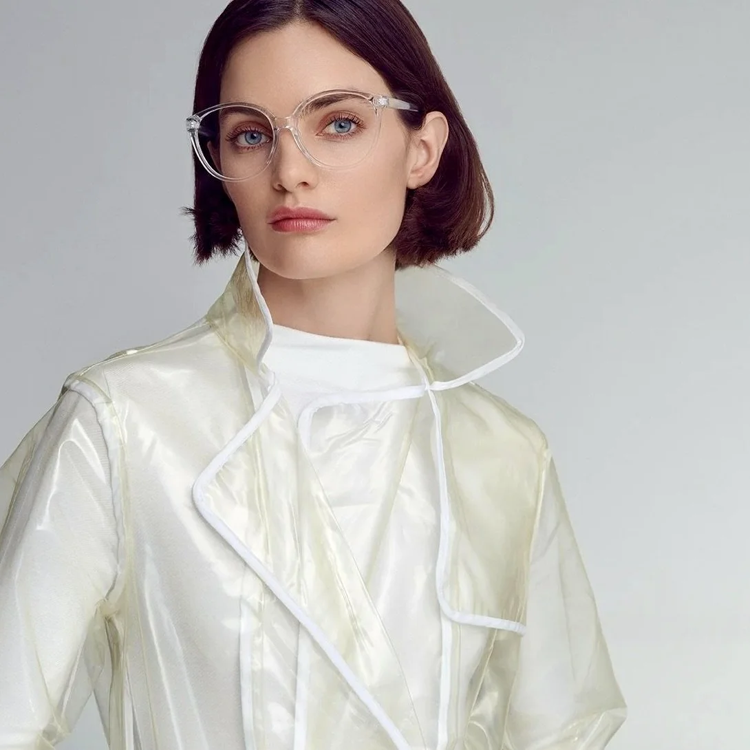 A woman with short brown hair wearing clear glasses and a white, shiny coat against a plain background.