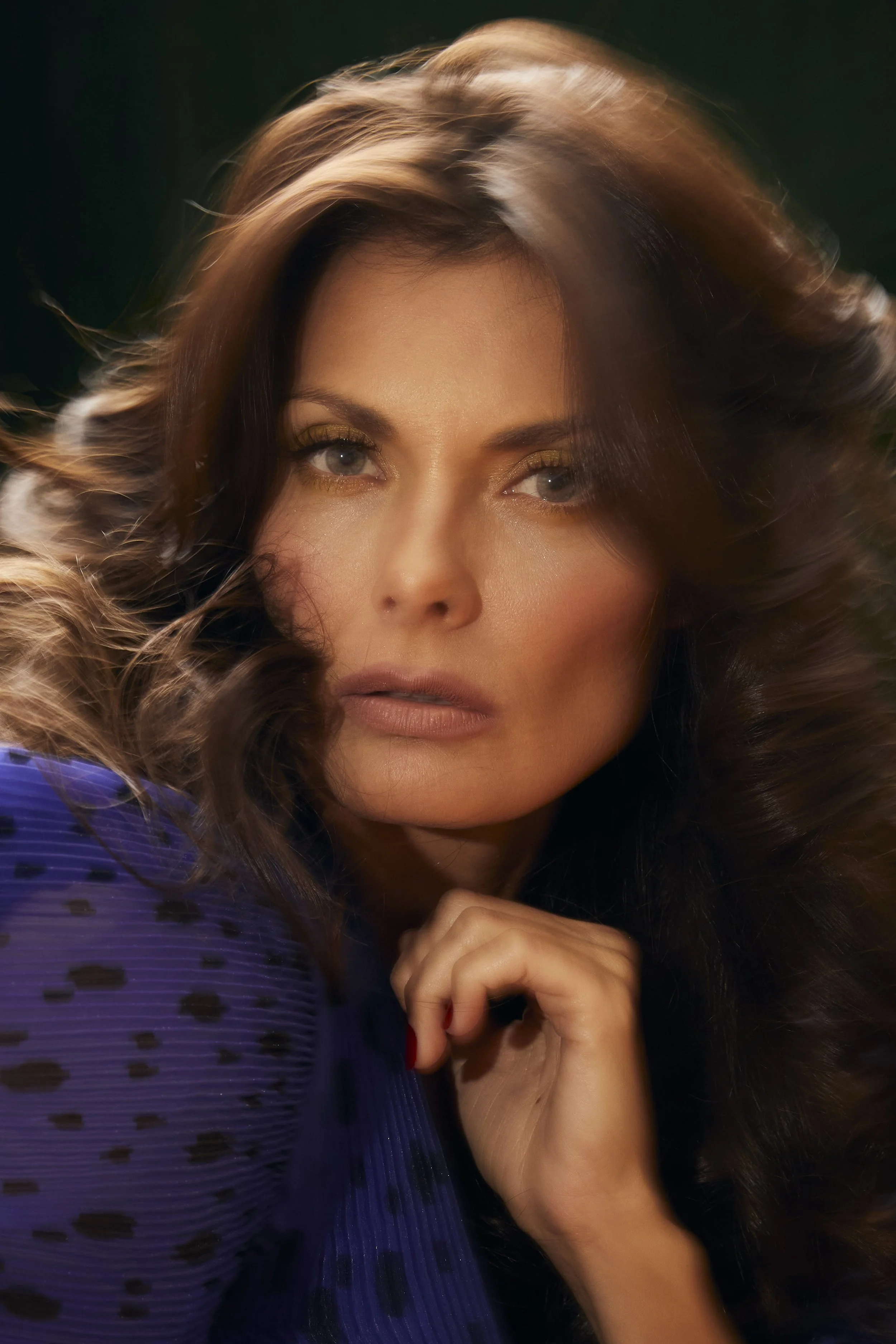 A close-up portrait of a woman with wavy hair and makeup, wearing a purple dress with black dots, resting her chin on her hand.