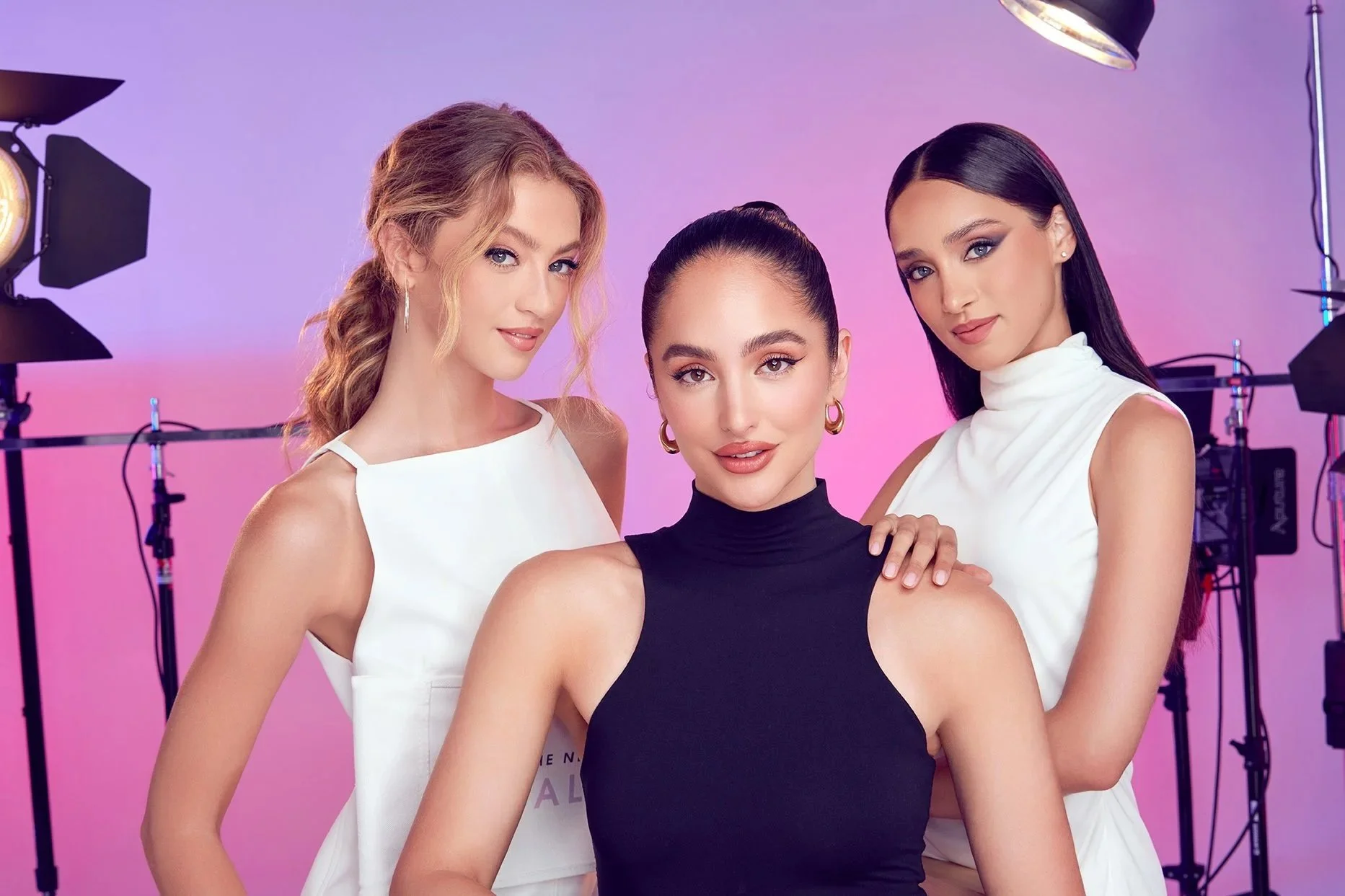 Three women posing together in a studio with professional lighting equipment, purple and pink background, dressed in elegant attire.
