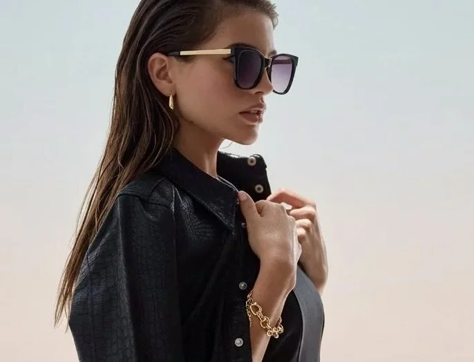 A woman wearing large black sunglasses, gold earrings, a gold bracelet, a black leather jacket, and a black top, standing outdoors against a plain sky background.