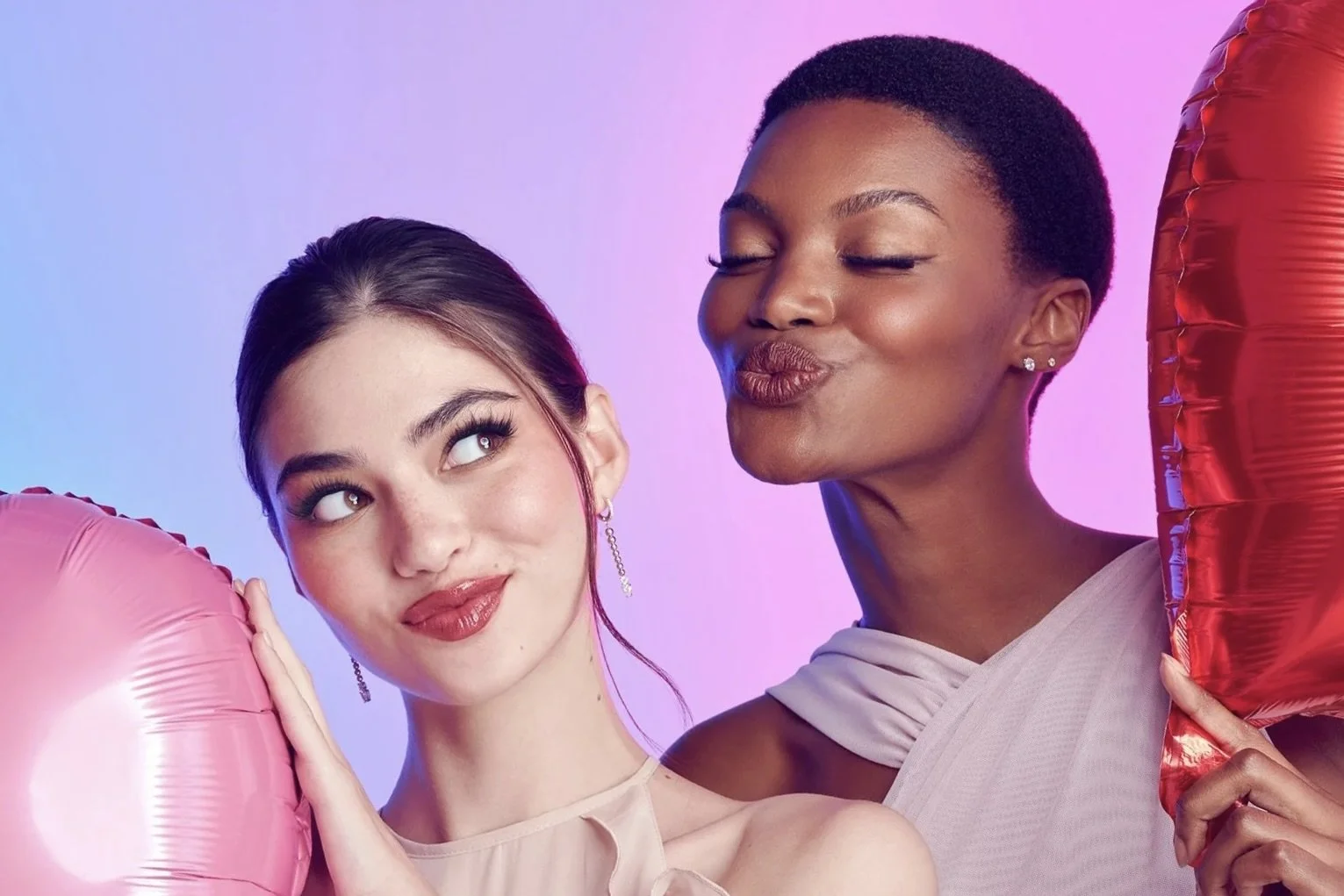 Two women holding pink and red balloons in a studio with a purple and blue background, making playful expressions.