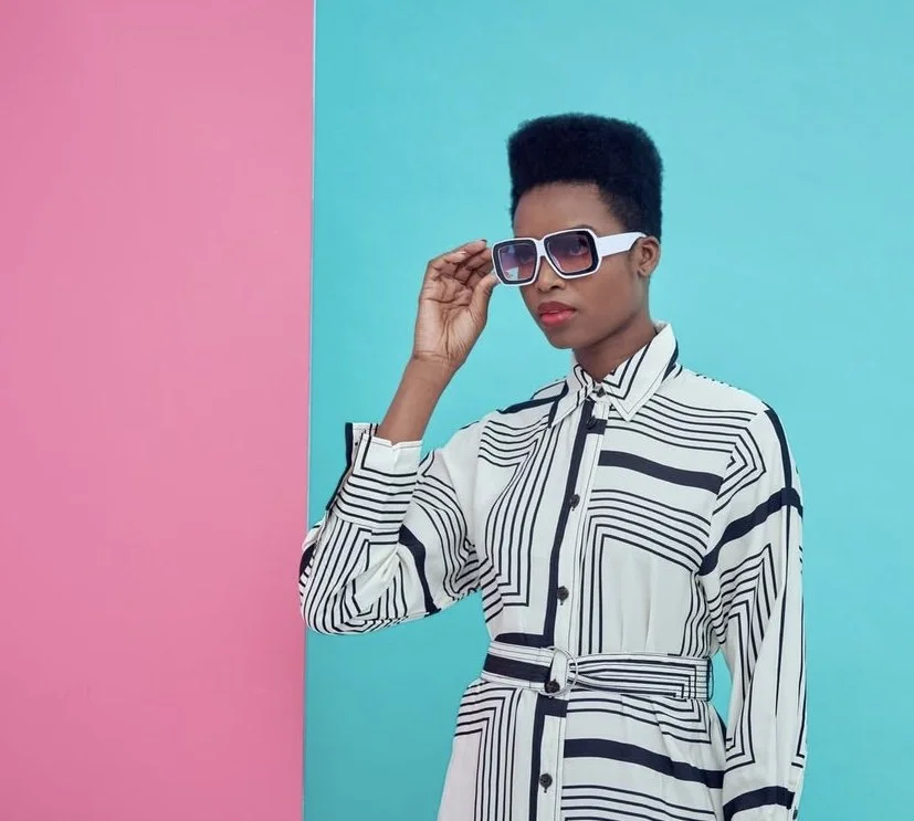 A woman with dark skin and a high flat-top hairstyle wearing large sunglasses and a black-and-white patterned dress standing against a split pink and blue background.