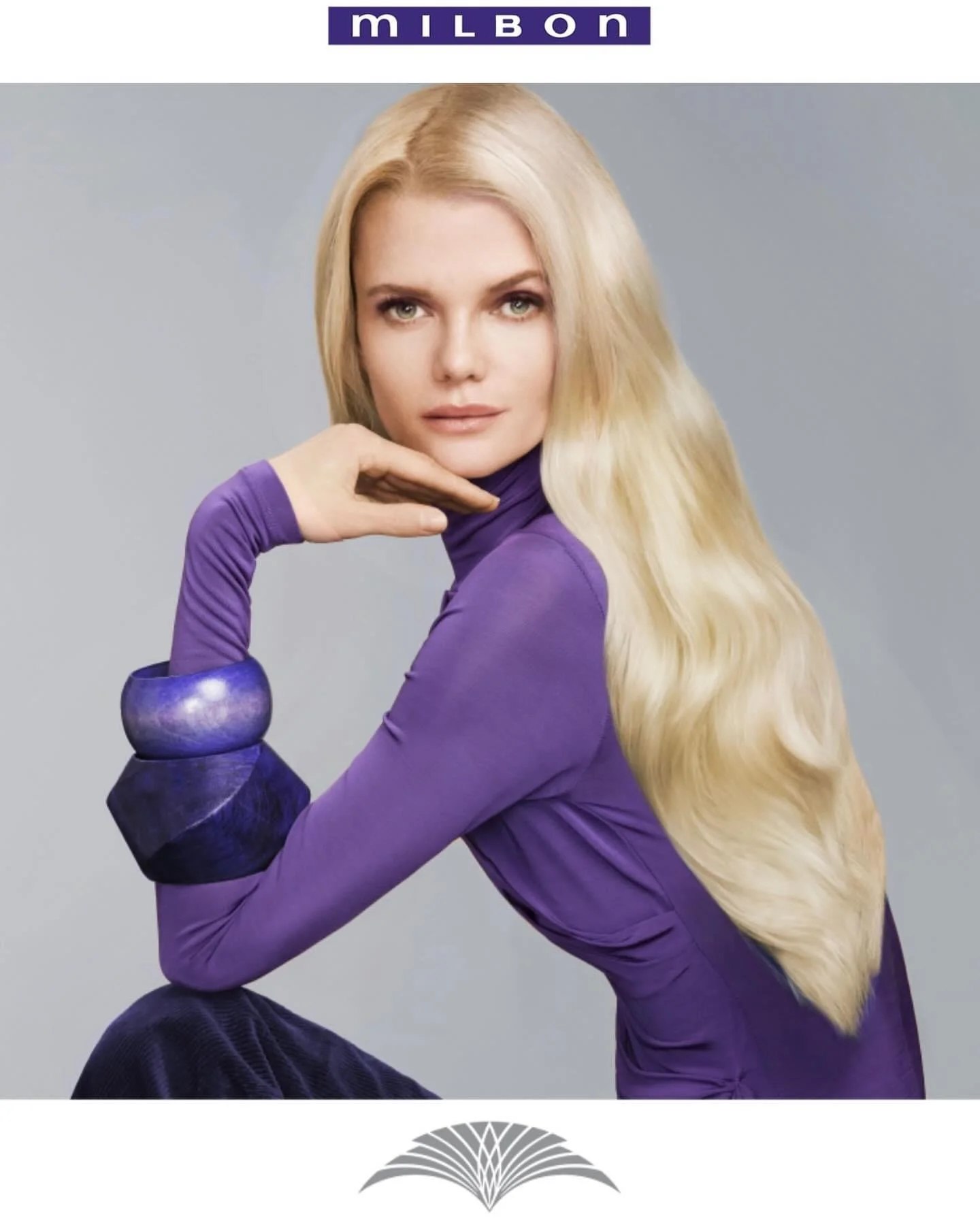 A woman with long blonde hair styled in loose waves, wearing a purple long-sleeve top with black accents on the sleeves, posing against a light gray background. She is resting her chin on her right hand, with her elbow on her knee, looking directly a