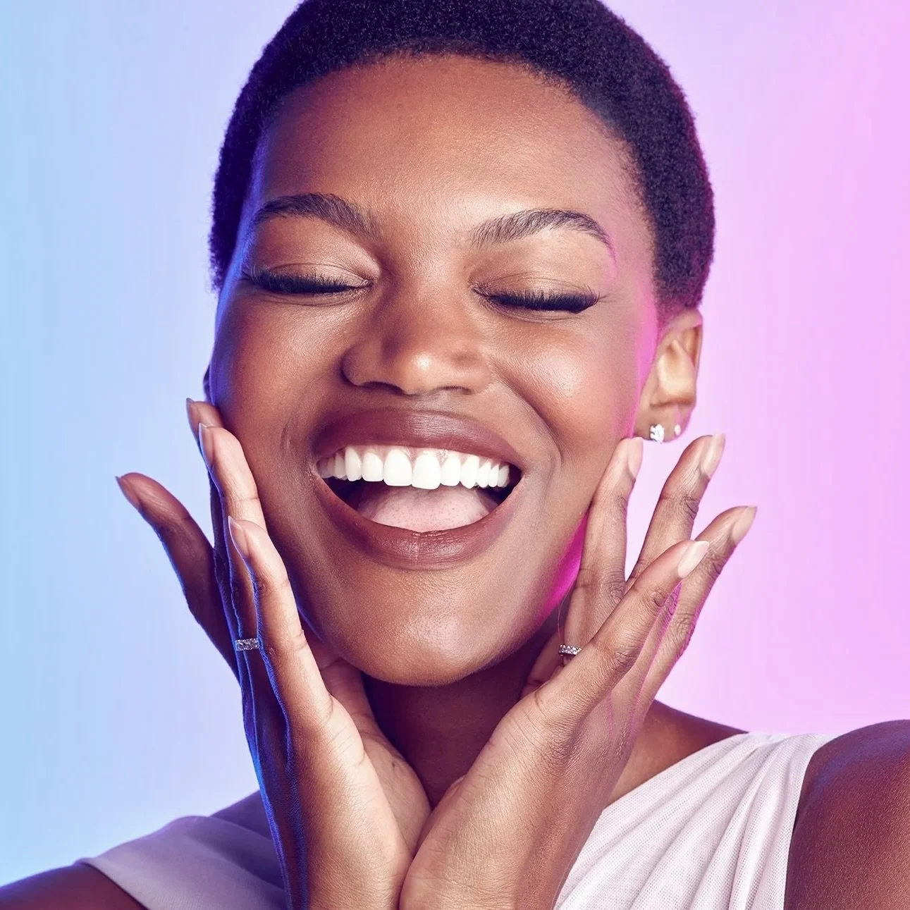 A woman with short hair smiling with eyes closed, showing teeth, holding her face with both hands, with a background of gradient purple and blue colors.