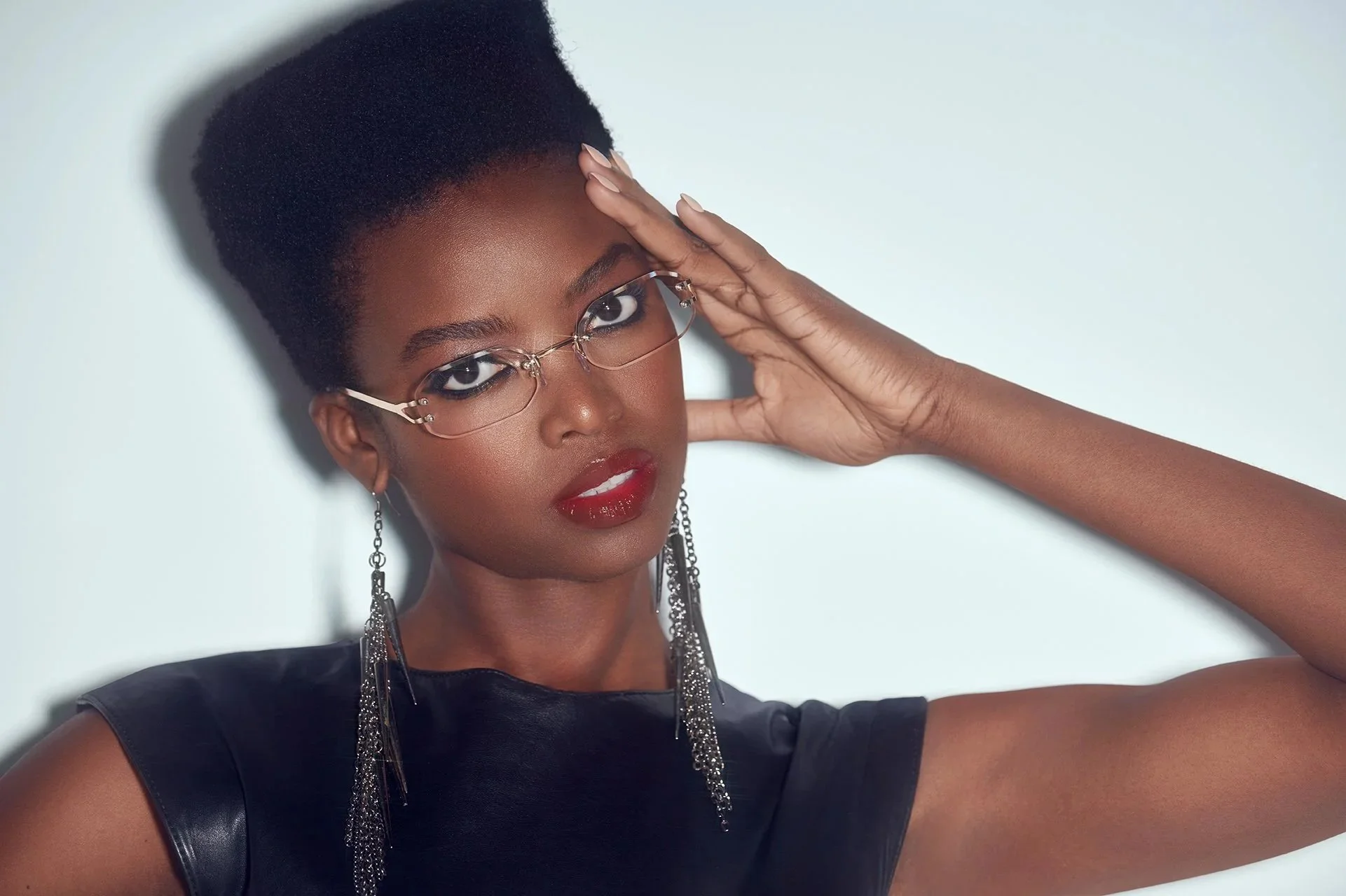 A woman with dark skin, short natural hair, wearing glasses, bold red lipstick, and long earrings, looking directly at the camera with her hand near her forehead against a plain light background.