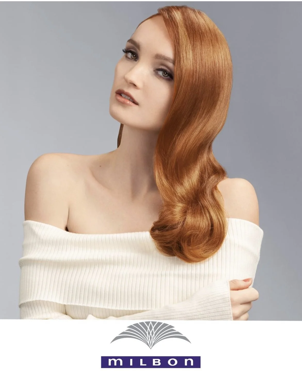 A woman with long, shiny, wavy red hair, wearing an off-the-shoulder white sweater, looking at the camera with a slight smile against a gray background.