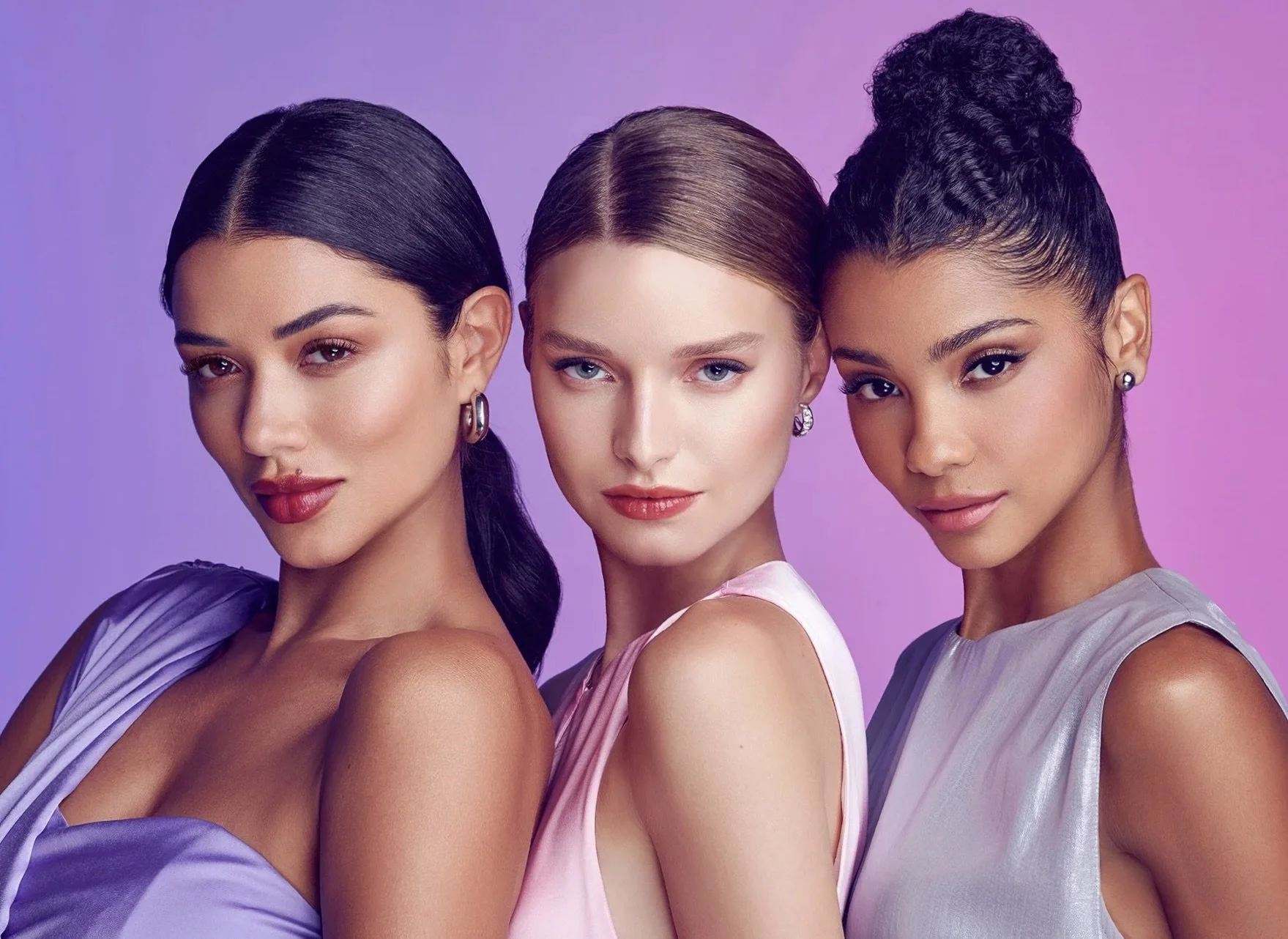 Three women with different skin tones and dark, light, and curly hair, wearing pastel-colored dresses, posing closely against a pink and purple gradient background.