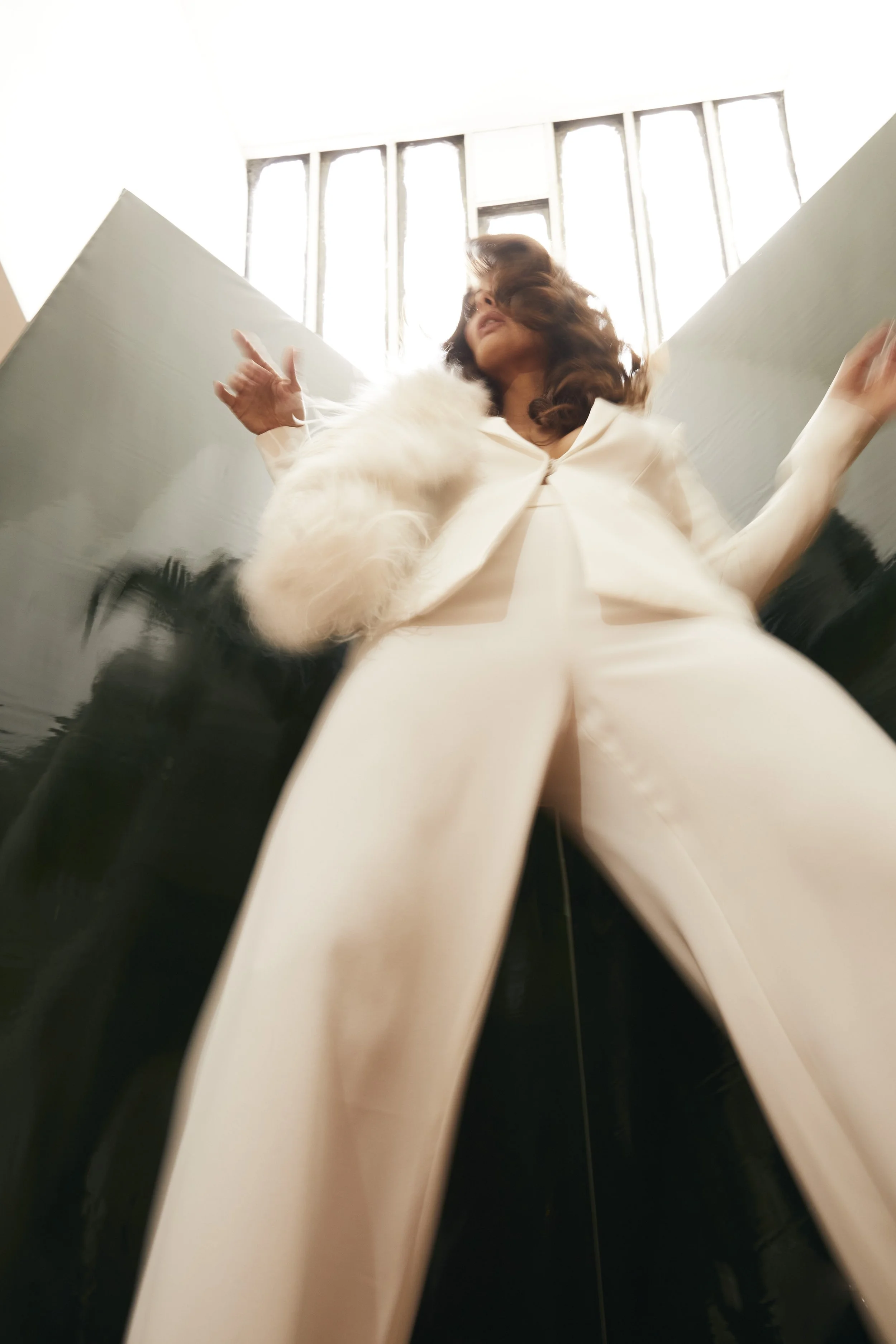 A woman with brown hair, wearing a white outfit with a fur collar, is seen from below with her face partially obscured, standing in front of large windows with sunlight streaming through.