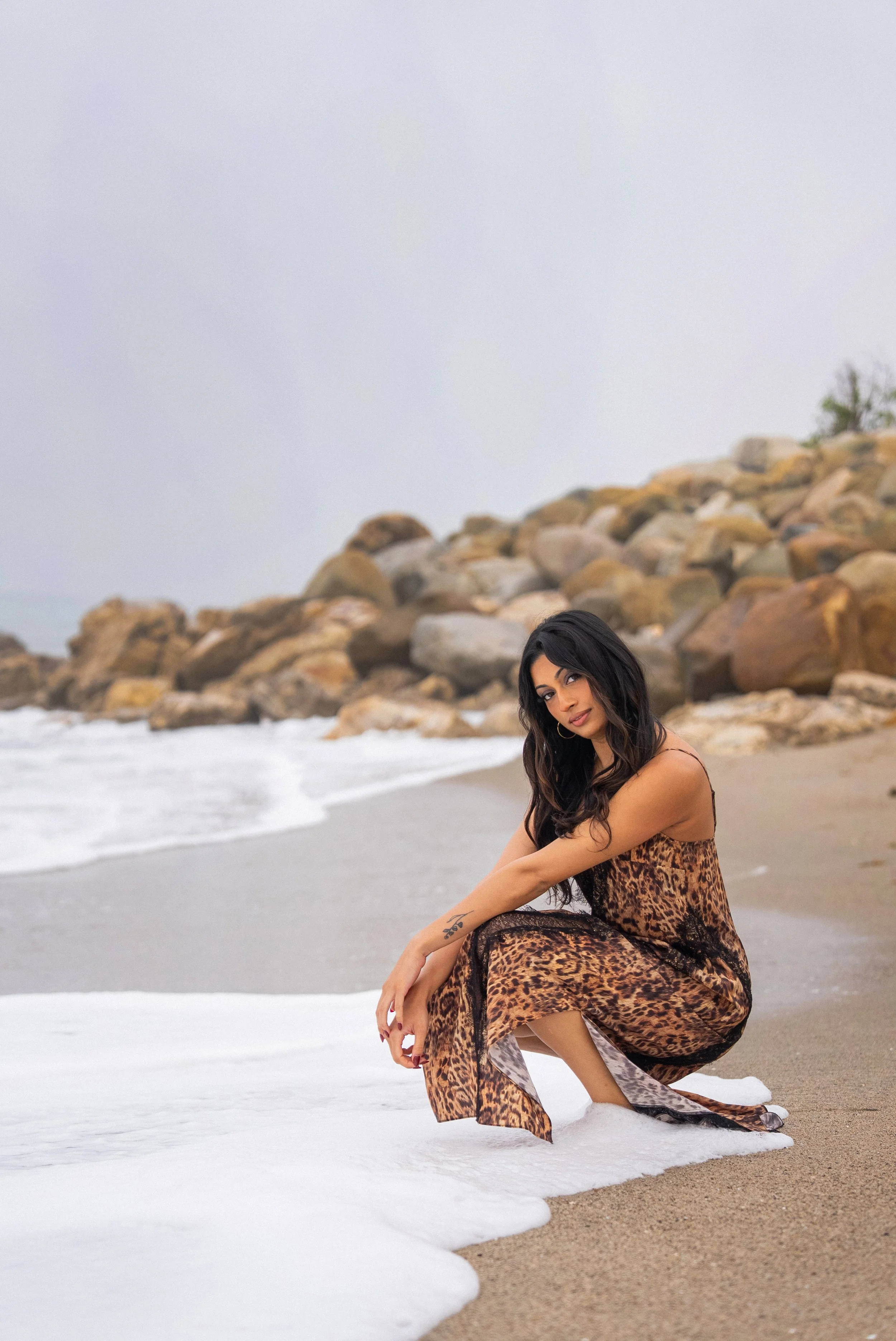 Woman in leopard print dress squatting on sandy beach with rocks and ocean waves in background