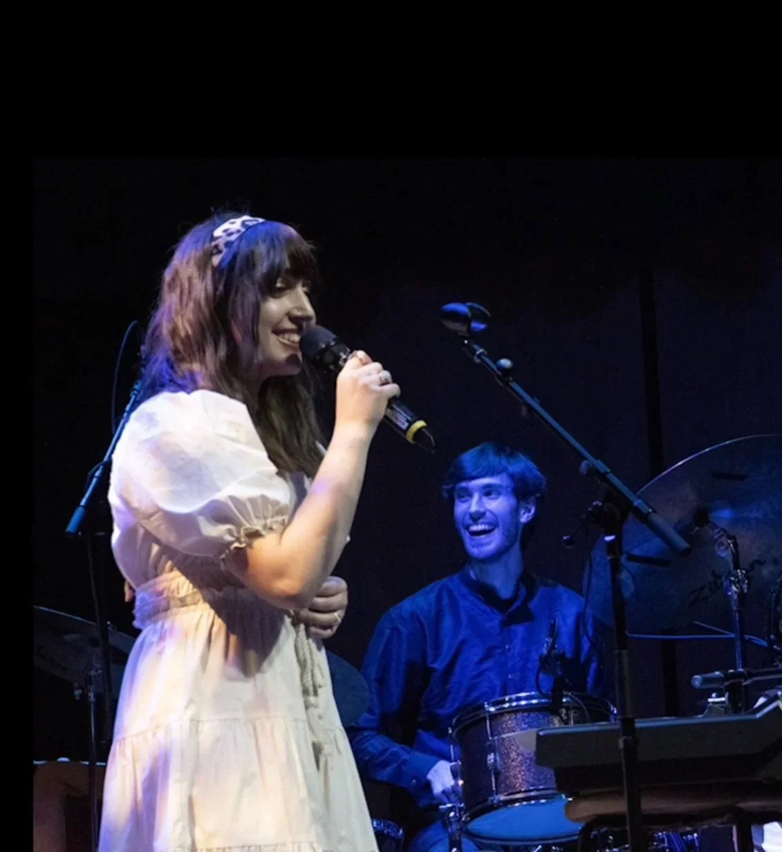 A woman with shoulder-length hair, wearing a white dress and a blue headband, singing into a microphone on stage. She is smiling and standing next to a man with dark hair, playing percussion instruments, who is smiling and looking at her. The background is dark with stage equipment.