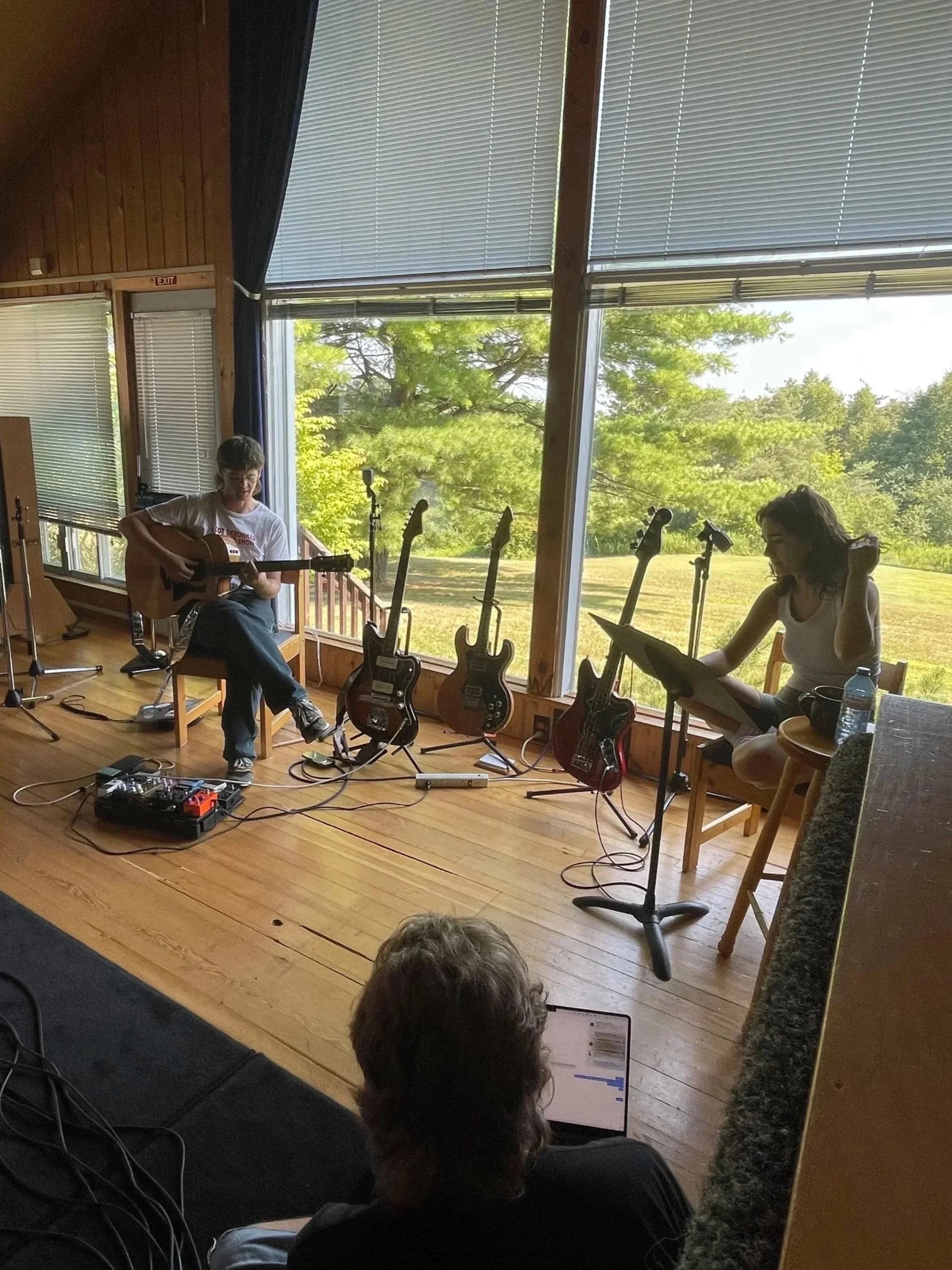 A man playing electric guitar and a woman reading music in a room with large windows and several electric guitars on stands outside, with a person sitting on the floor controlling sound equipment.