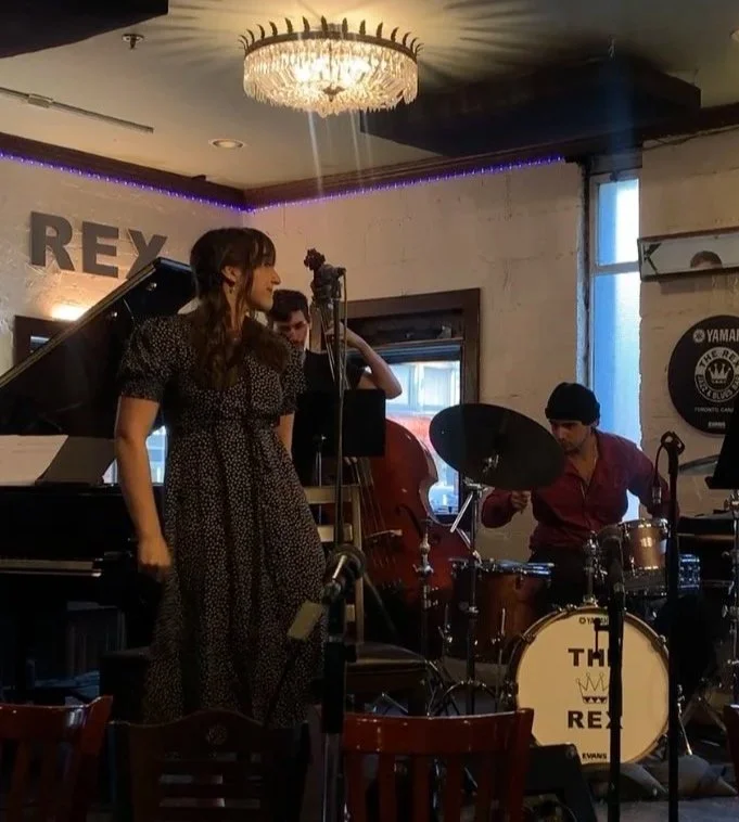 A woman singing at a jazz club stage with a pianist, double bassist, and drummer, under a chandelier with a sign reading 'REX' in the background.