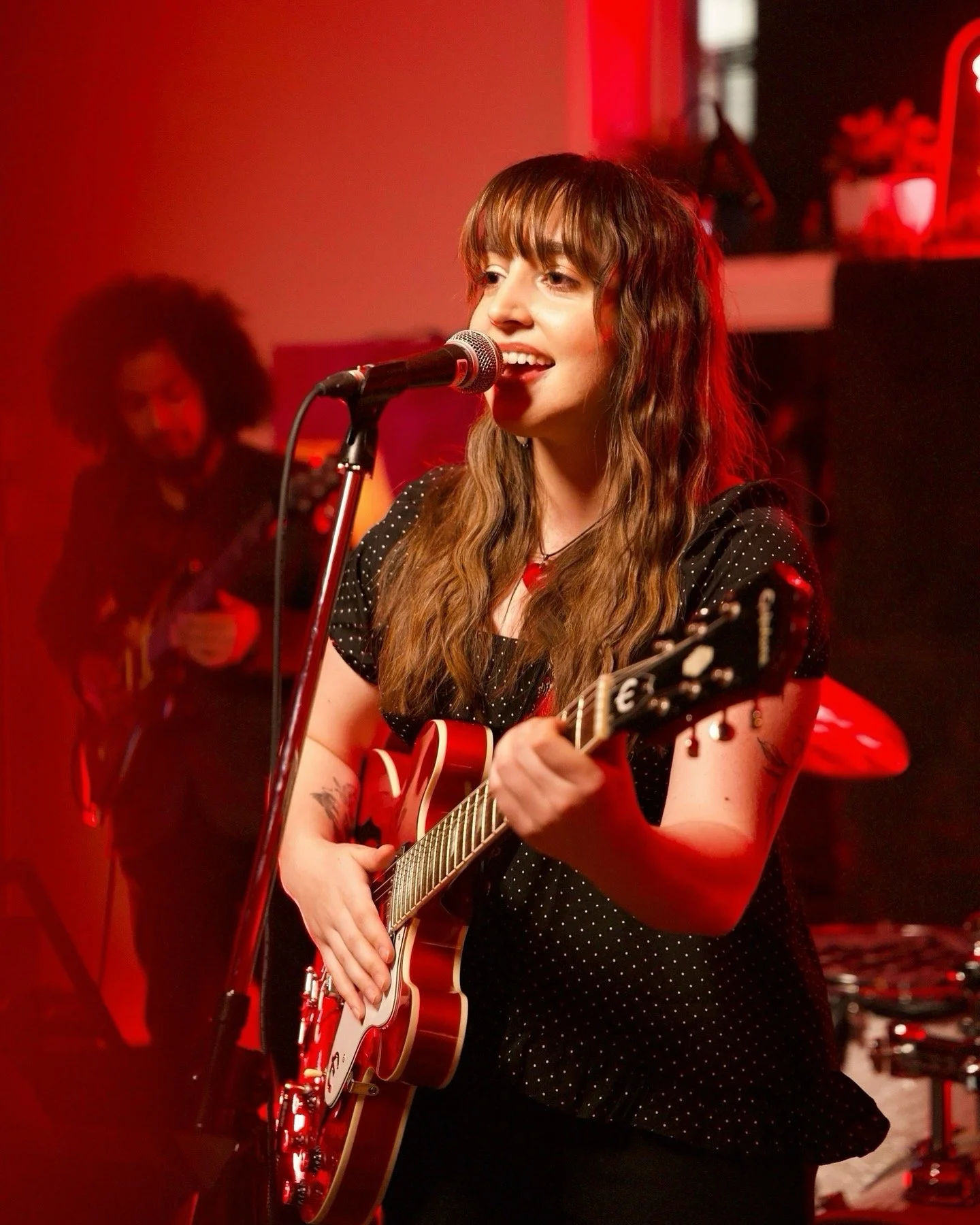 A woman with long wavy brown hair singing into a microphone while playing a red electric guitar on stage with a red and black background.