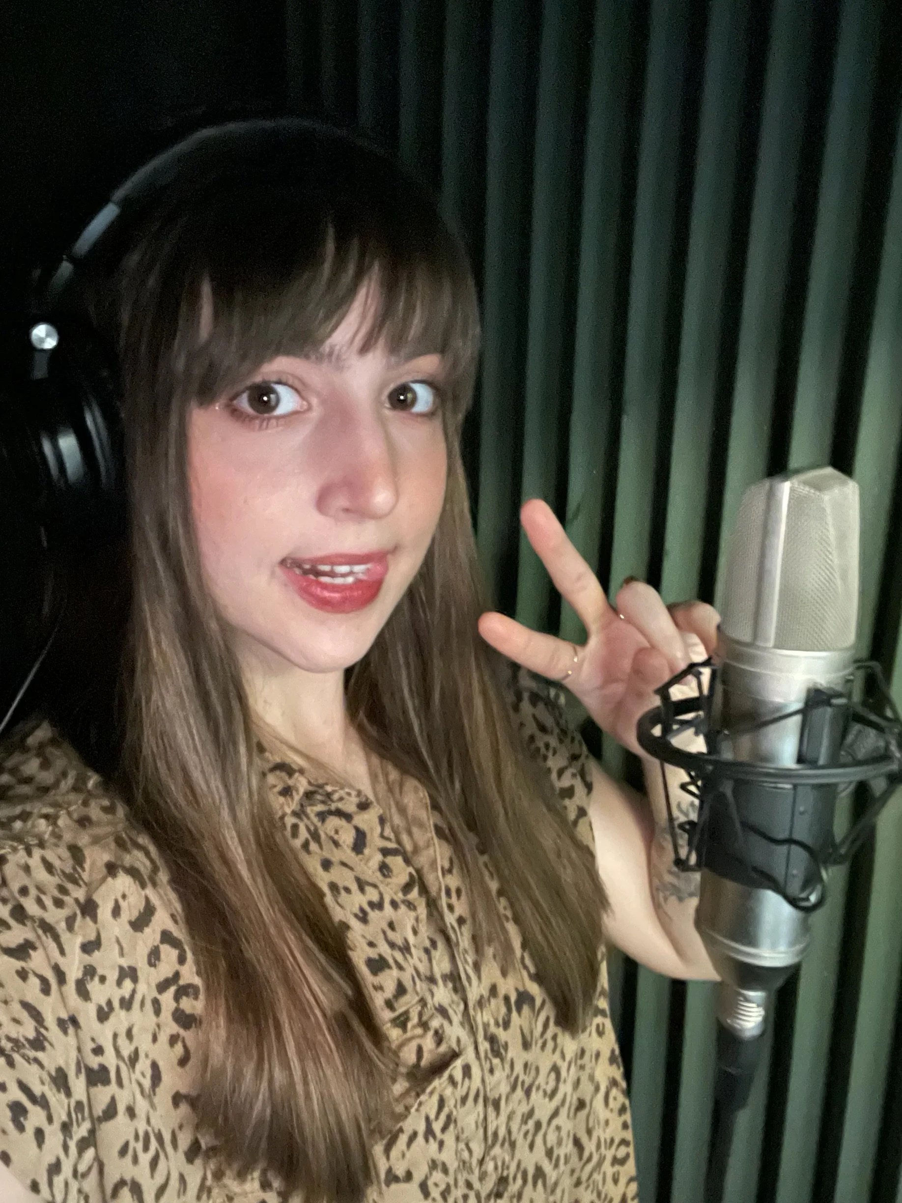A young woman with long brown hair and bangs, wearing a leopard print shirt, standing in a recording studio with a large microphone and green acoustic panels in the background. She is wearing headphones, smiling, and making a peace sign with her right hand.
