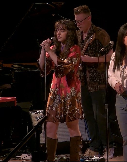 A young woman singing into a microphone on stage, with a man playing guitar behind her, and part of another person visible on the right.