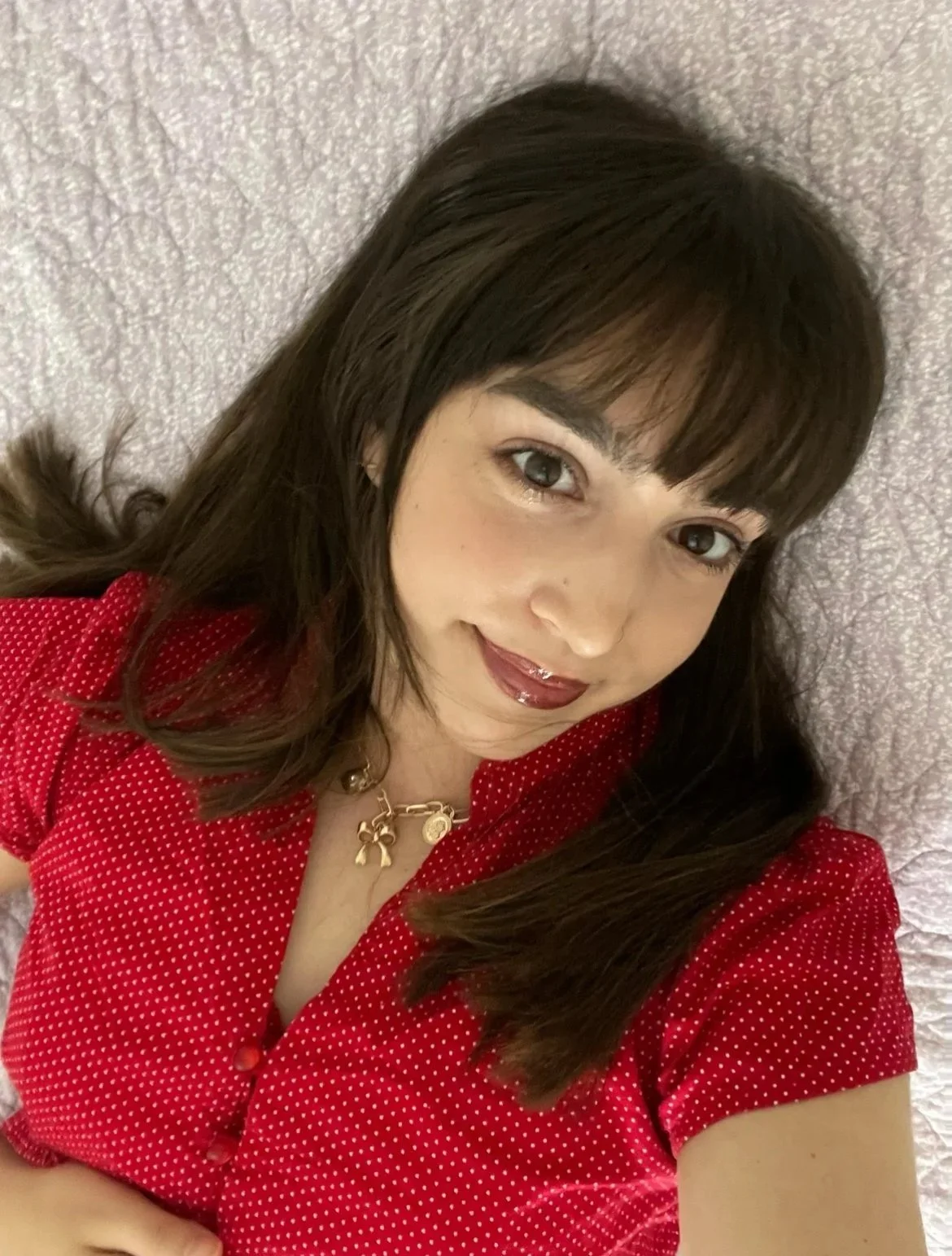 Close-up selfie of a woman with dark brown hair, wearing a red polka dot blouse and a gold necklace, lying on a lilac textured duvet.