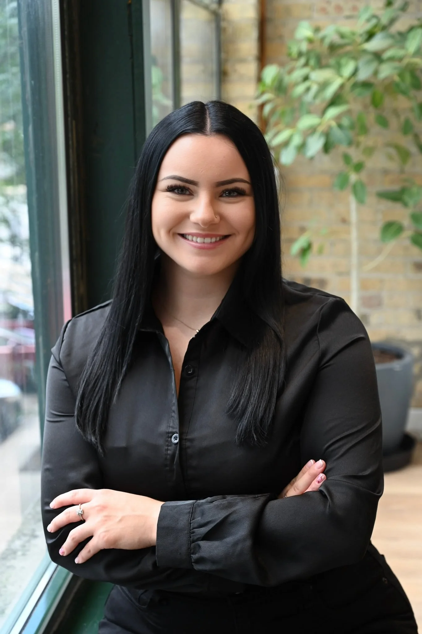 A woman with long black hair smiling, wearing a black button-up shirt, standing with her arms crossed indoors next to a large window with a brick wall and indoor plants in the background.