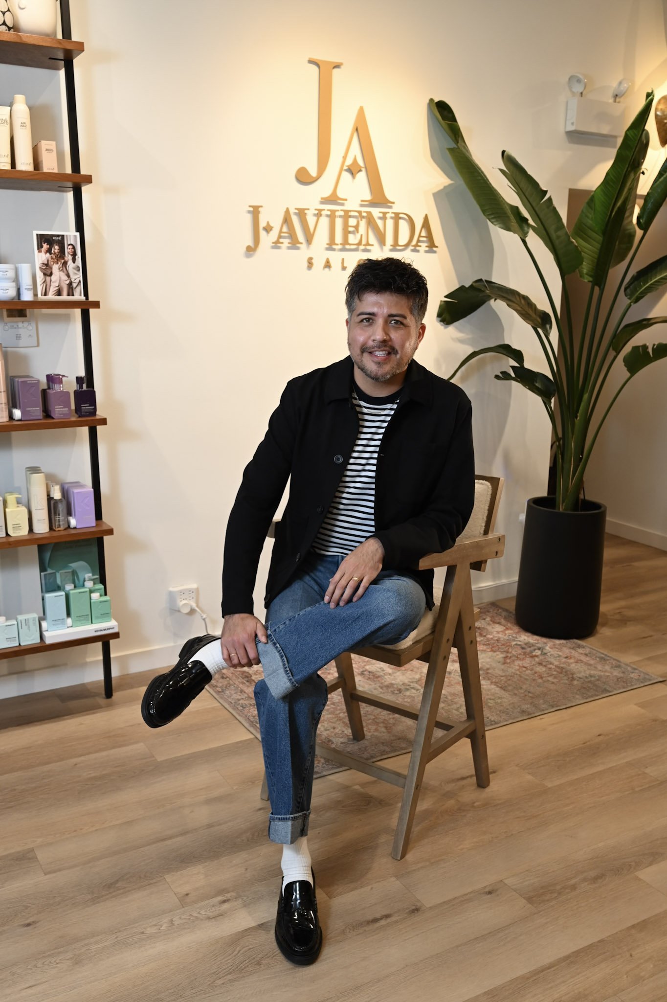 A man seated on a wooden chair inside a salon, with a large plant and shelves of hair products behind him, and the salon's logo on the wall reading 'J A Javienda Salon'.