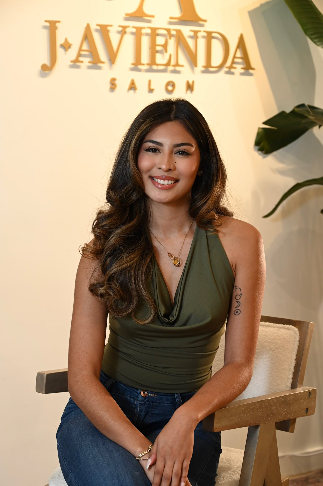 A woman with long, wavy brown hair and tan skin, sitting on a chair at a salon, smiling at the camera. She is wearing an olive-green sleeveless top and blue jeans, with a necklace and a bracelet. Behind her is a sign that reads 'JAVENDA SALON' on a light-colored wall, with a large green plant partially visible on her right side.