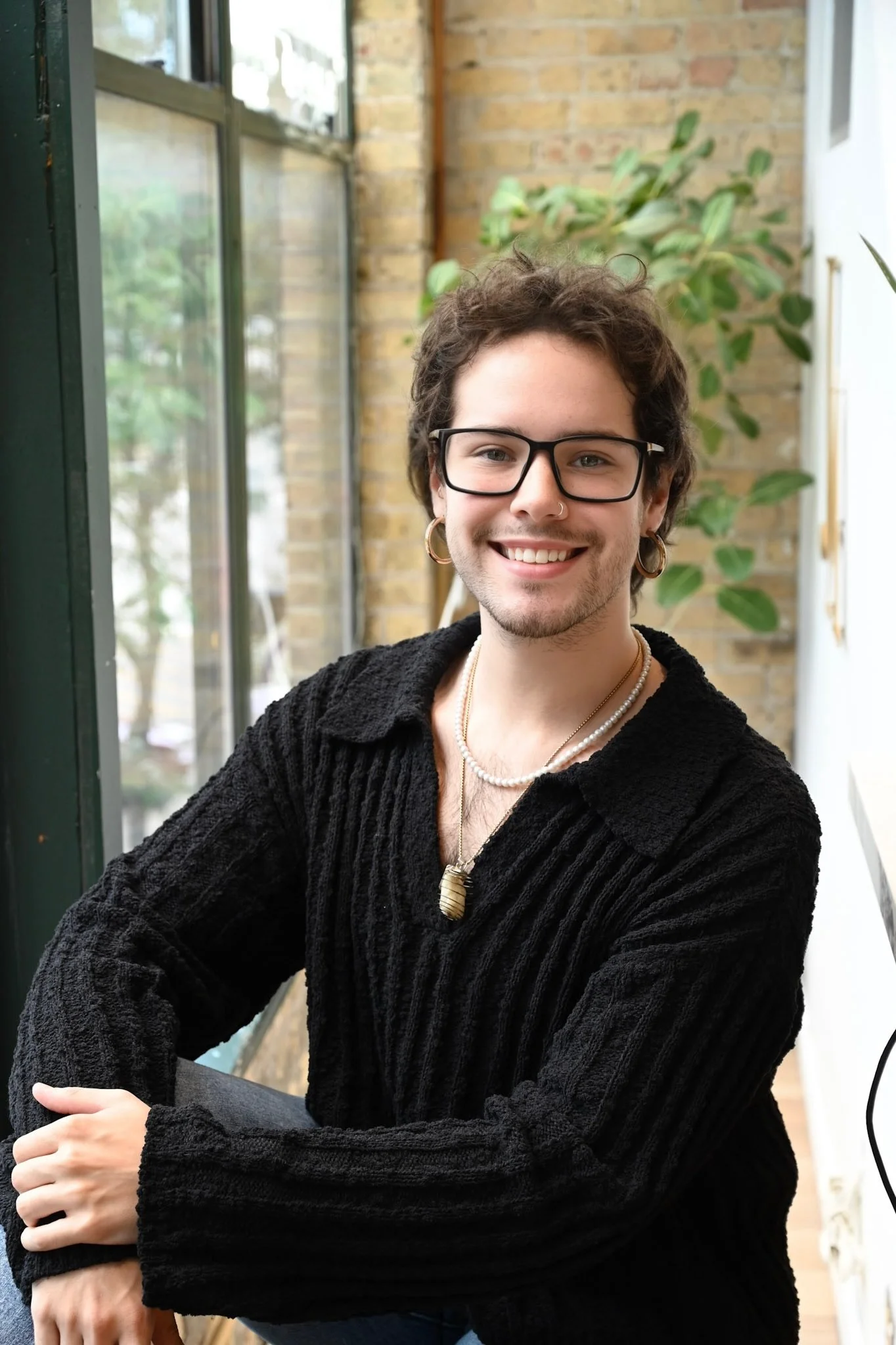 A young person with short curly hair, glasses, hoop earrings, and a nose ring smiling at the camera. They are wearing a black knit sweater, layered necklaces, and are seated by a window with a plant and brick wall in the background.