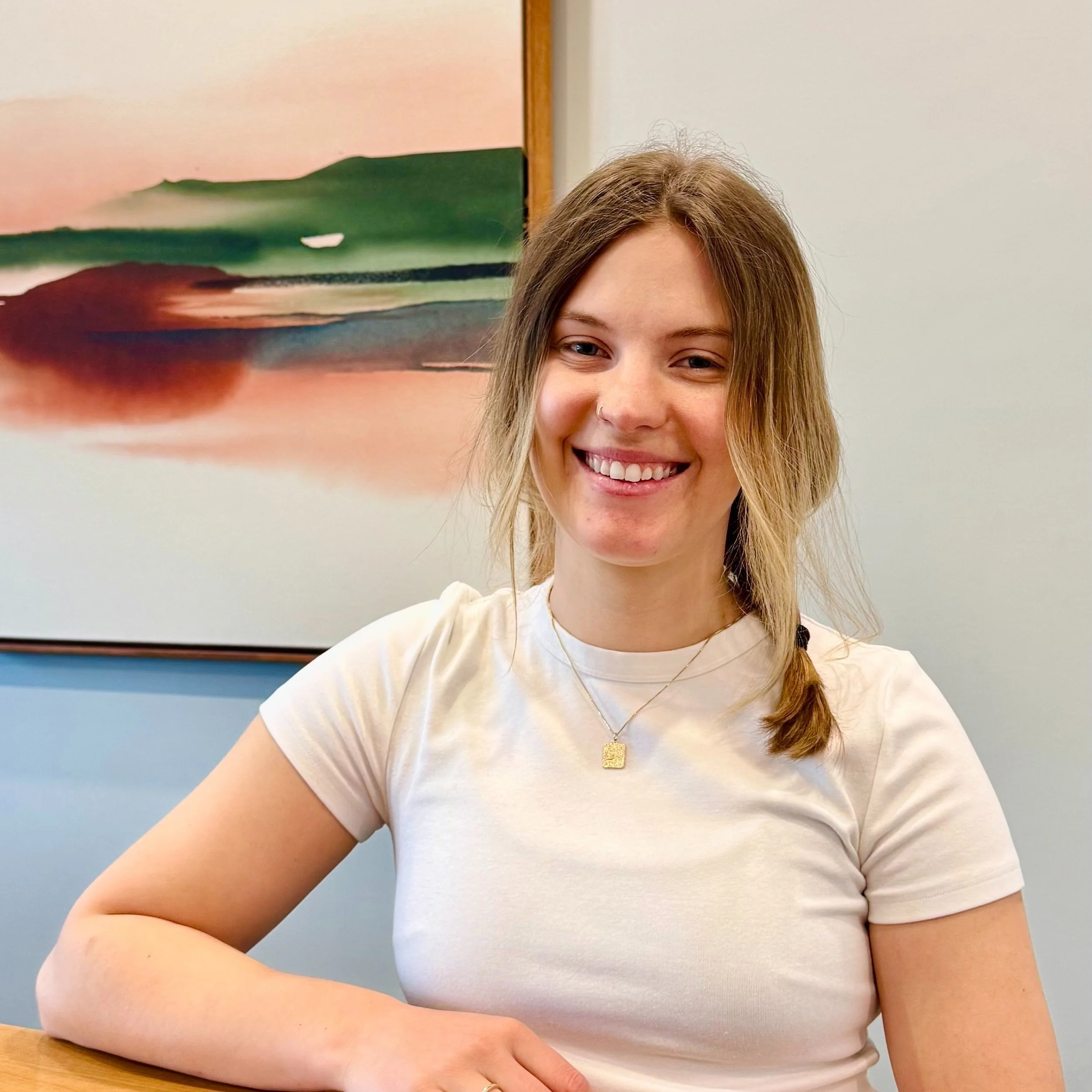 A young woman with long, light brown hair styled in loose waves and a side braid, smiling while seated indoors. She is wearing a white t-shirt and layered necklaces, with a framed abstract landscape painting in the background.