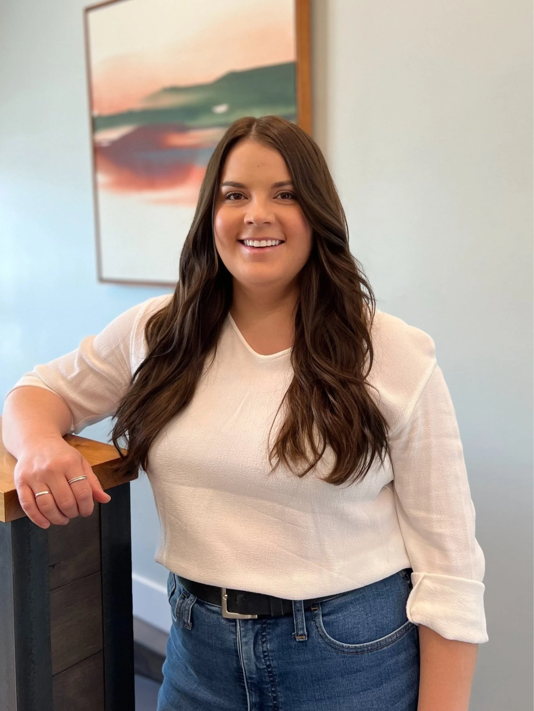 Receptionist with long brown hair wearing a white top and jeans, smiling while leaning on a wooden structure, with an abstract painting in the background.