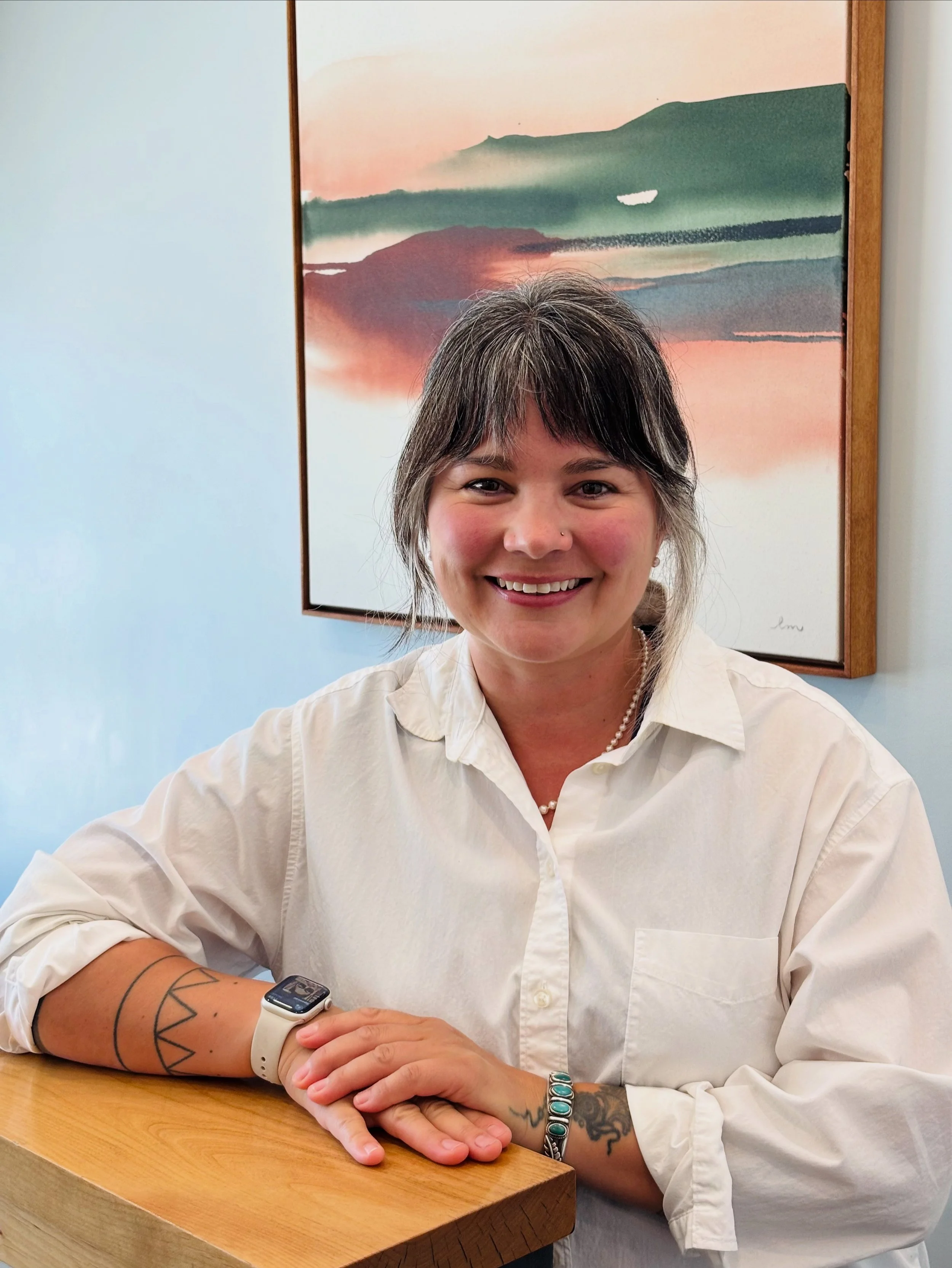 A woman with shoulder-length dark hair and a smile, wearing a white shirt, sitting at a wooden table with her hands folded. She is wearing a smartwatch and some jewelry, with a tattoo on her left arm. In the background, there is a framed landscape painting of a mountain and water with a sunrise or sunset sky.