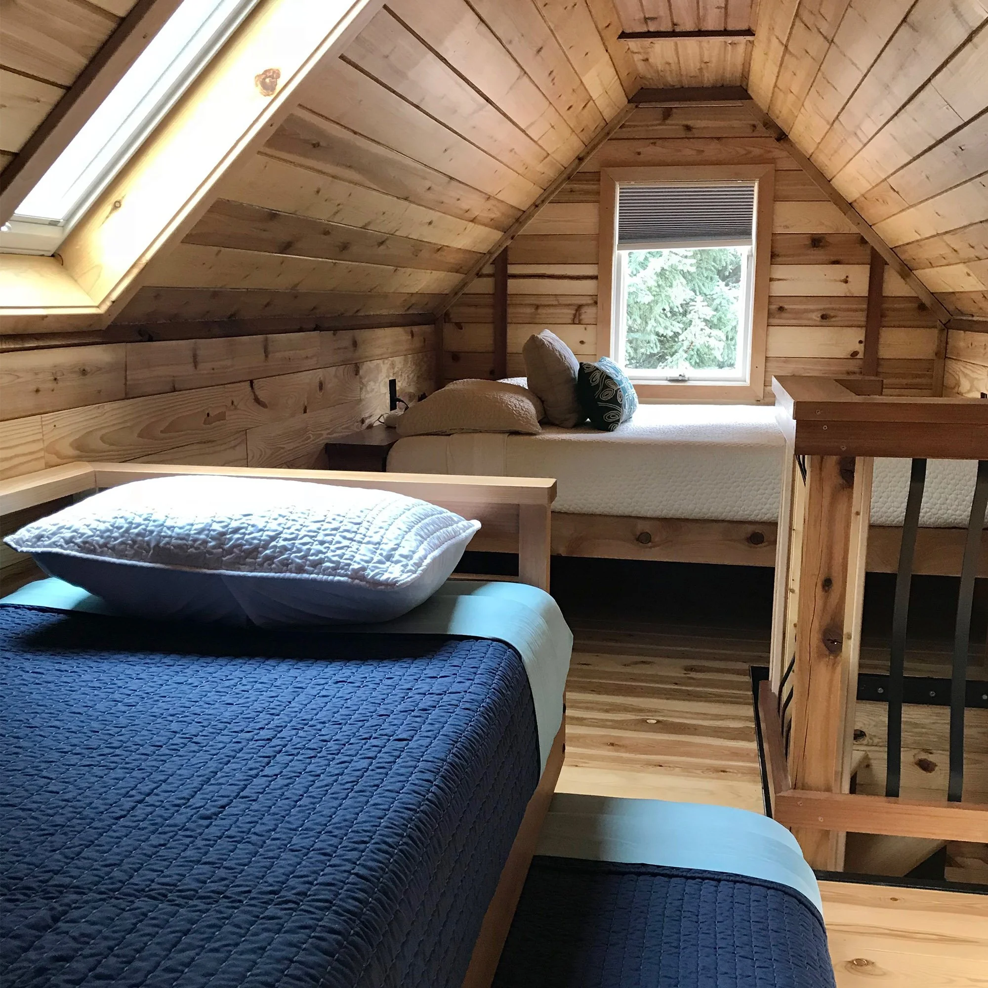 Cozy loft bedroom inside a tiny house at WeeCasa, featuring wood-paneled walls, a skylight, two neatly made beds with blue and white bedding, and a window overlooking trees.