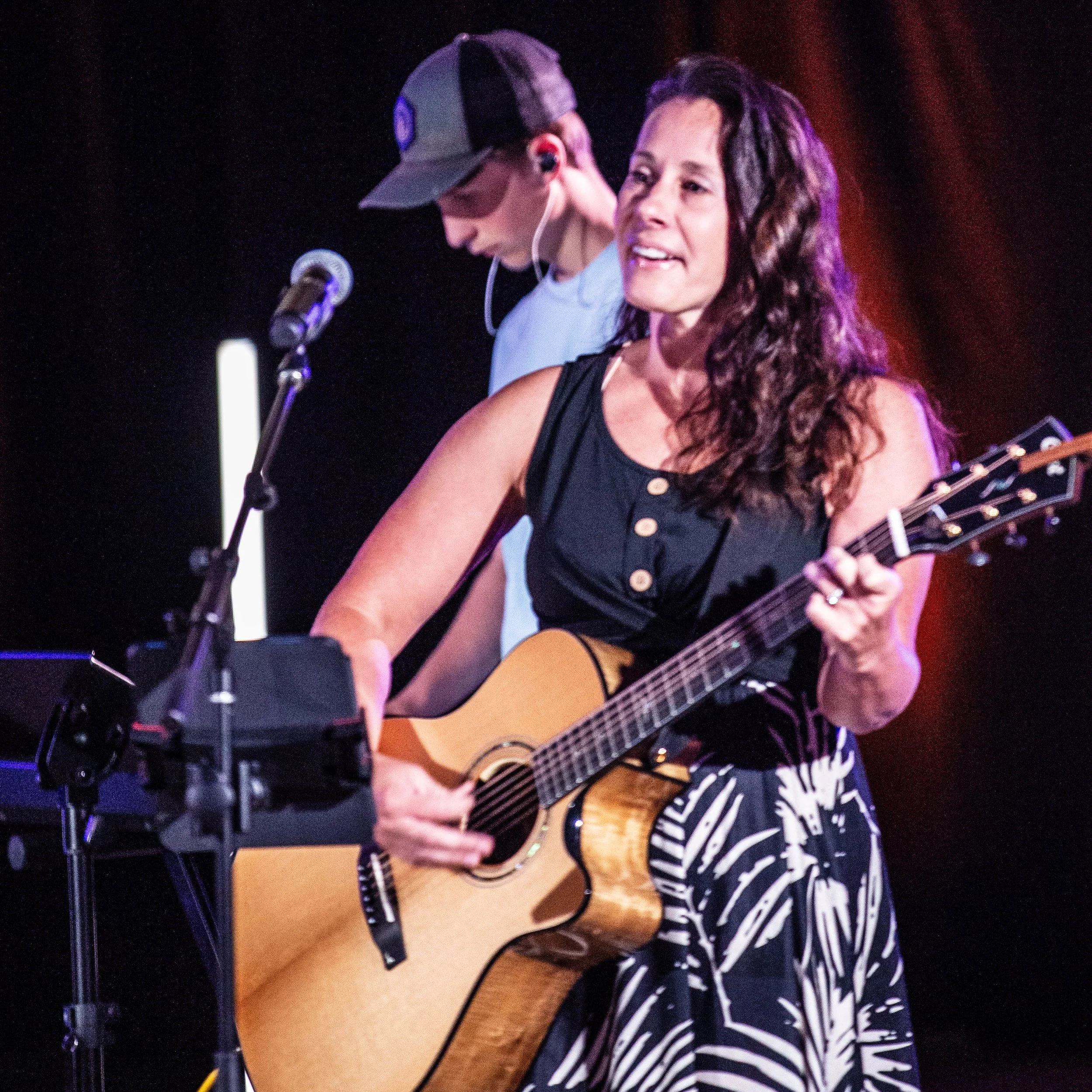 A woman playing an acoustic guitar on stage, with a man wearing a baseball cap and earpiece behind her, in a performance setting.