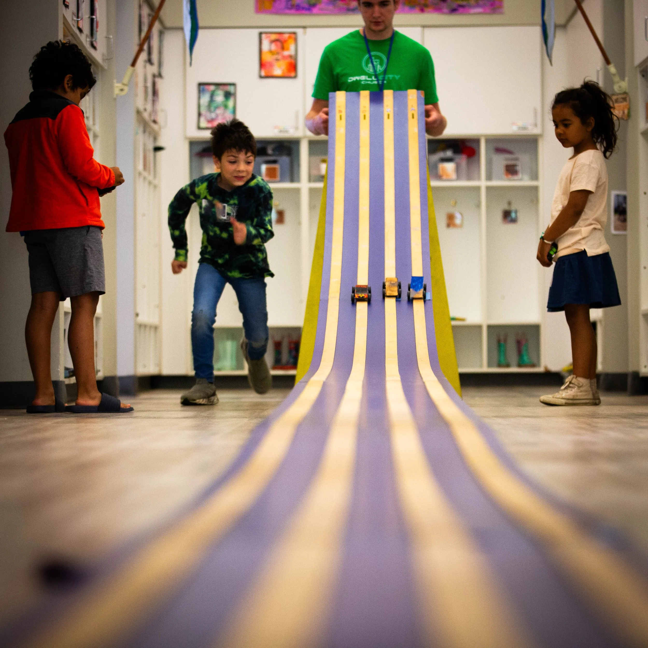 Children playing with mini race cars on a ramp in a classroom setting.