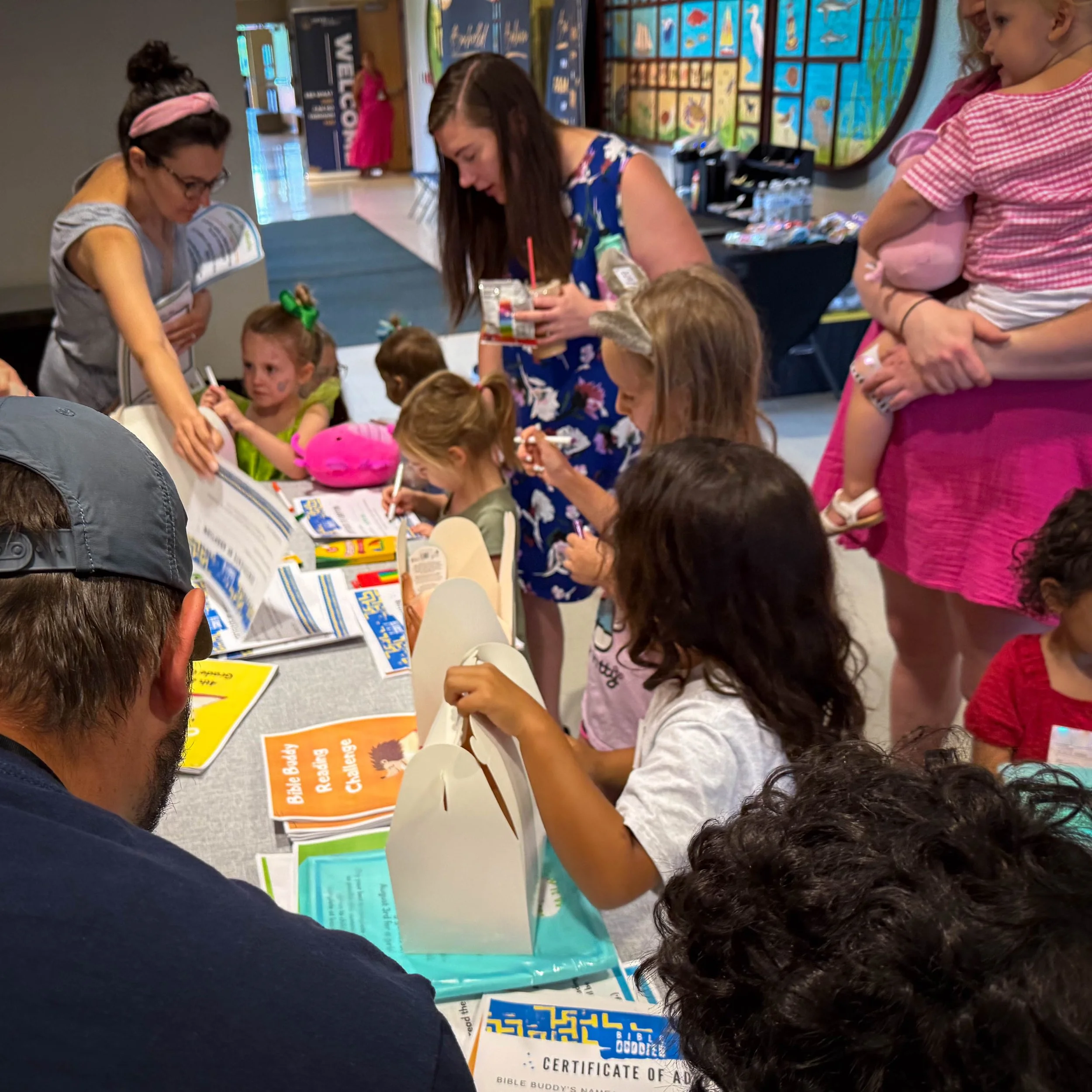 Children and adults gathered around a table signing books and holding supplies at a book signing event in a public space.