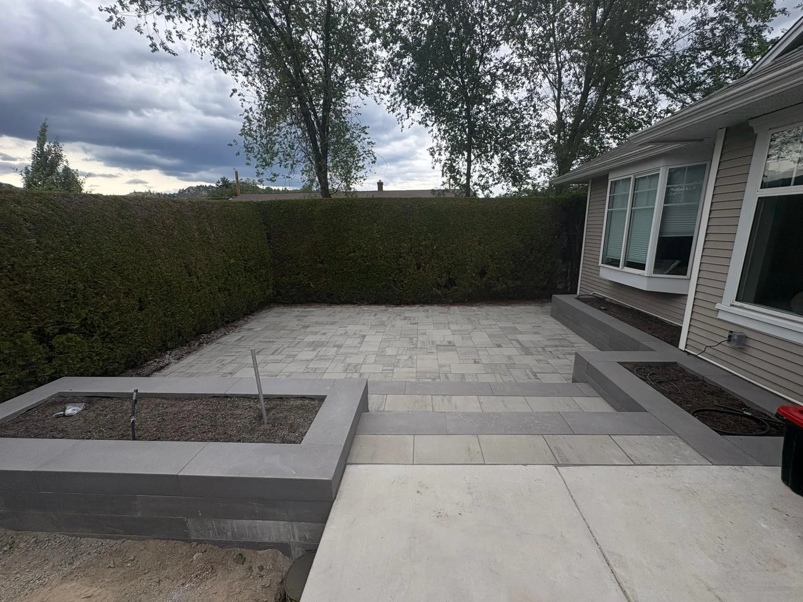 View of a backyard patio with newly installed gray paving stones, bordered by a house on the right and a tall hedge at the back, with a cloudy sky overhead.