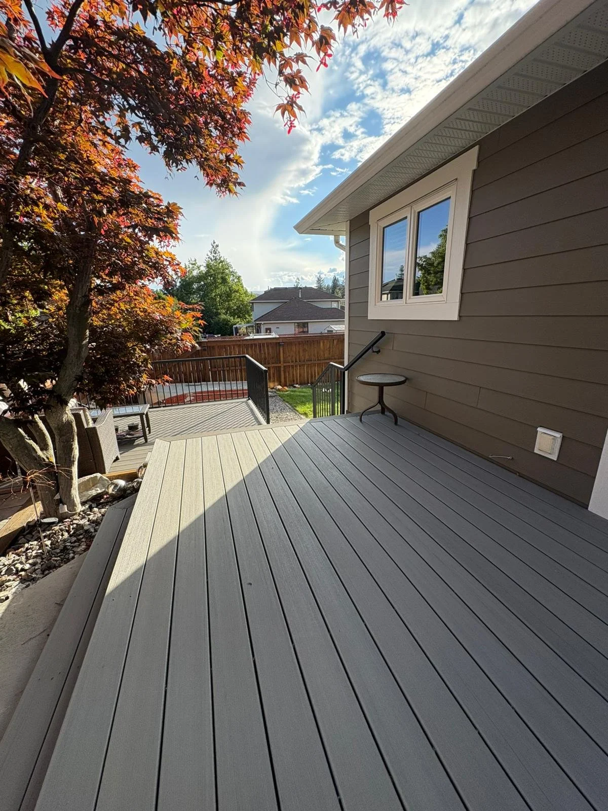 A wooden deck attached to a house with brown siding, a small round table, a tree with reddish leaves, and a view of neighboring houses under a partly cloudy sky.