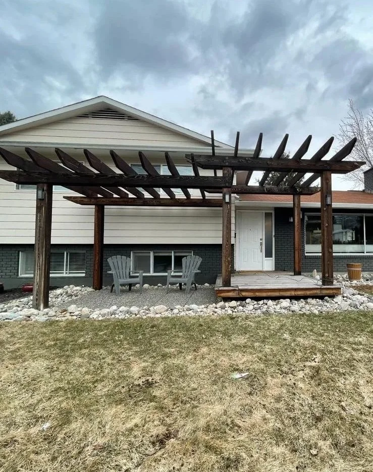 House with a wooden pergola on a small patio, two Adirondack chairs, gravel ground cover, and a grassy yard in the foreground.