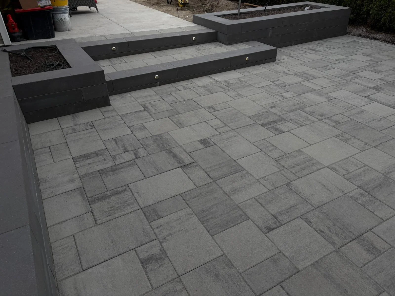 A paved outdoor area with grey stone tiles, a raised black stone planter, and steps with embedded lights.