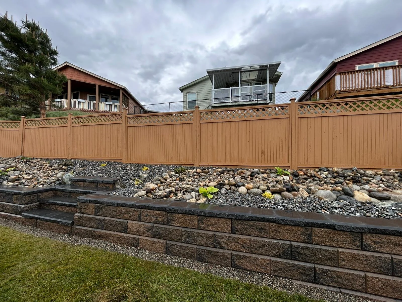 A backyard with a stone retaining wall, small stairs, and gravel bed with rocks and a few plants, enclosed by a wooden fence. In the background, there are three houses with decks and a cloudy sky.