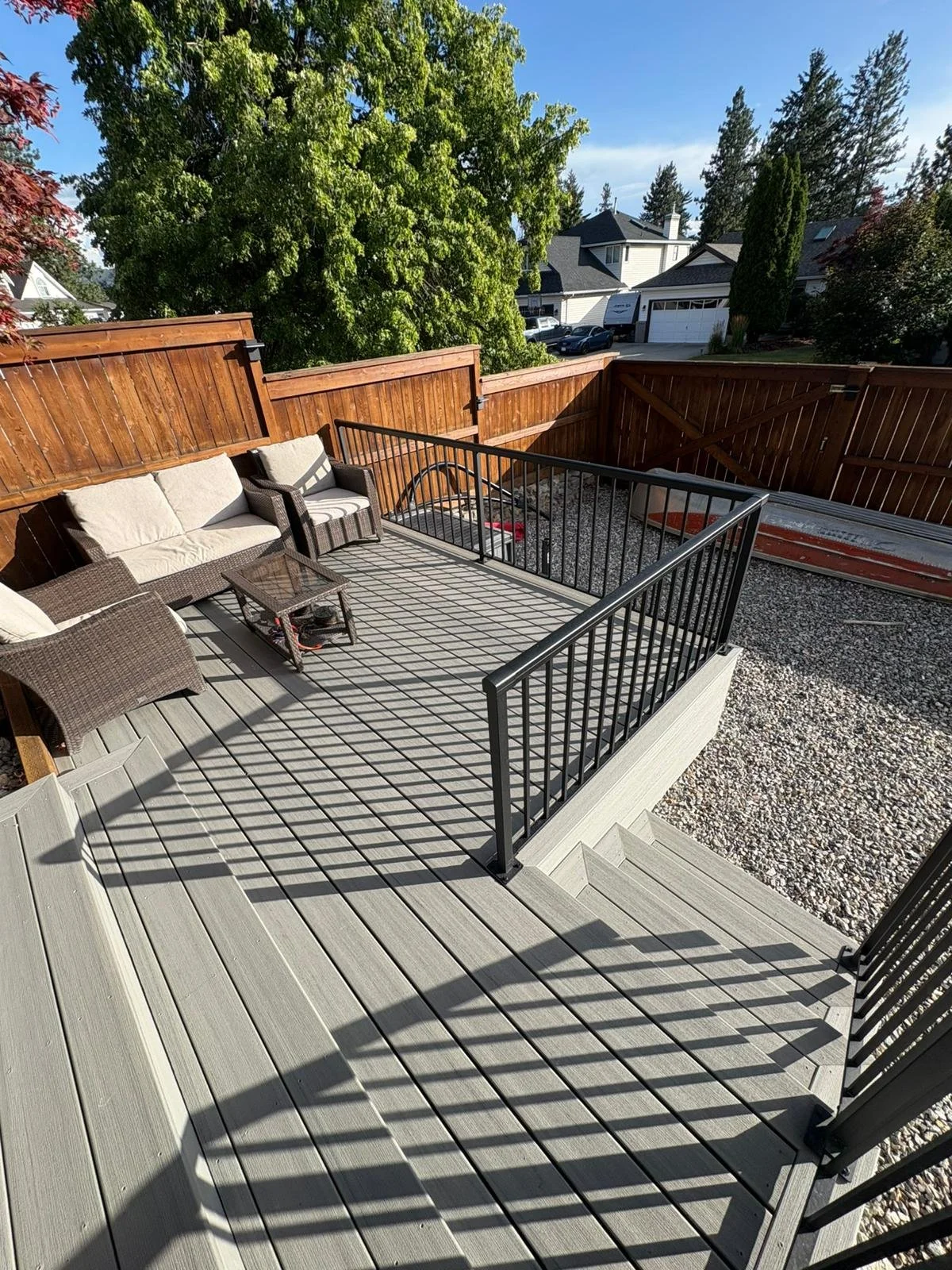 A backyard patio with wooden decking, black metal railing, outdoor furniture including a wicker sofa, two chairs, and a coffee table. There is a gravel area and a tall wooden fence surrounding the patio, with trees and houses visible in the backgroun