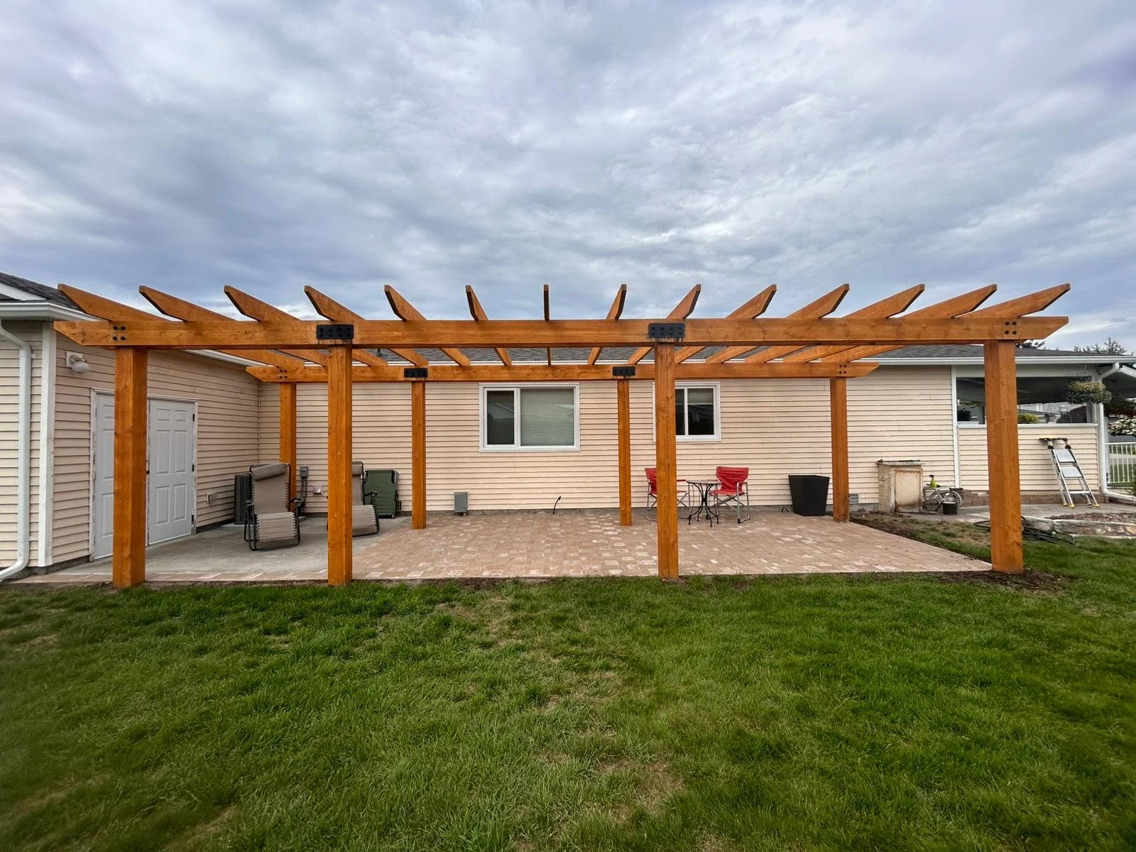 A backyard patio with a newly built wooden pergola over a brick-paved area, surrounded by green grass and a house with beige siding in the background.