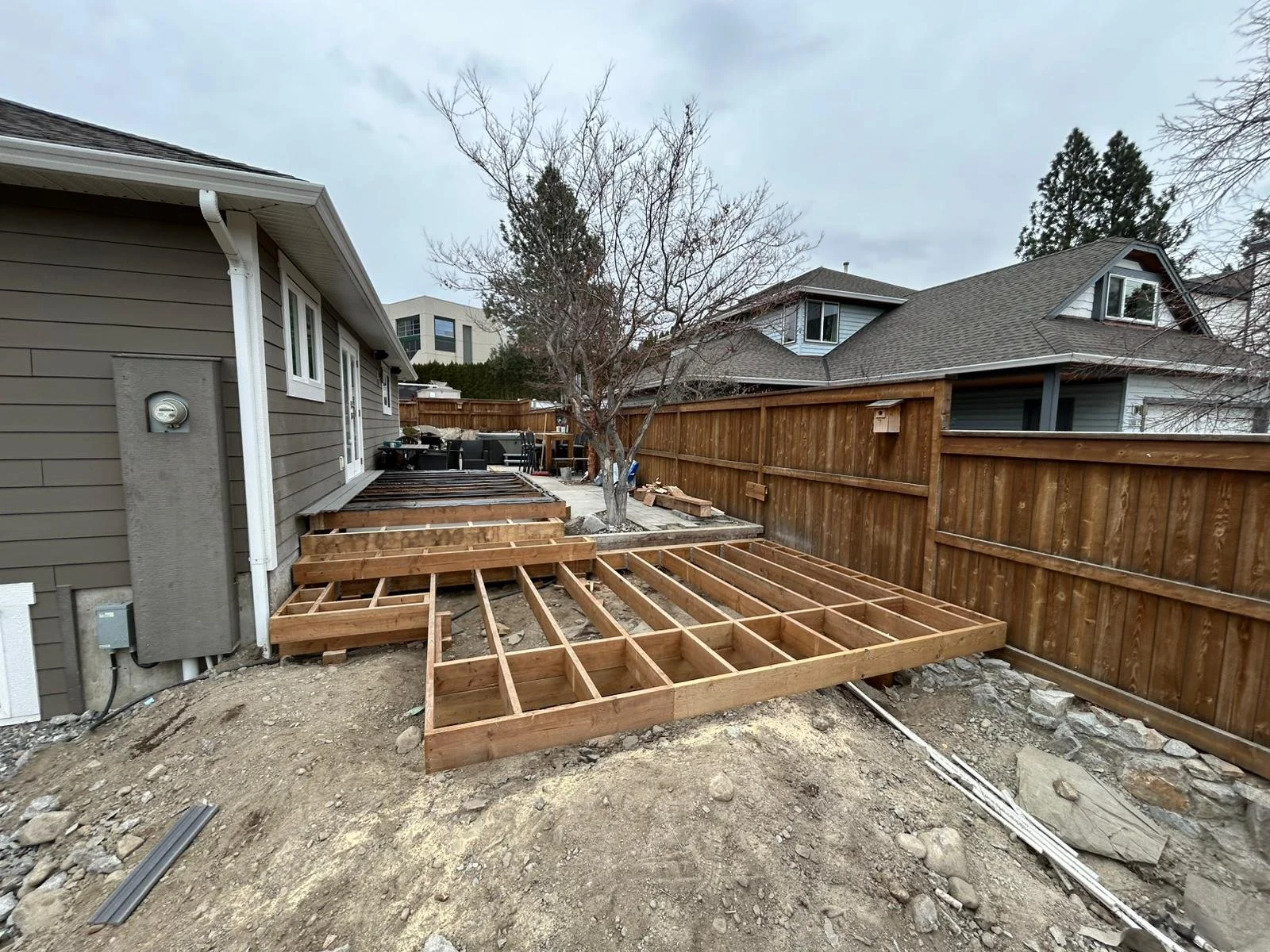 Wooden deck framing under construction in backyard, with neighboring houses and a wooden fence visible.