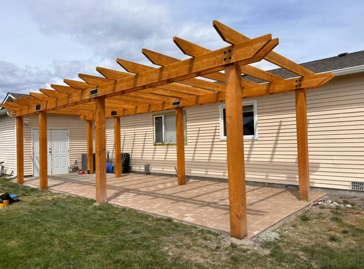 Backyard patio with a wooden pergola structure under construction, attached to a beige house with white trim and two windows.