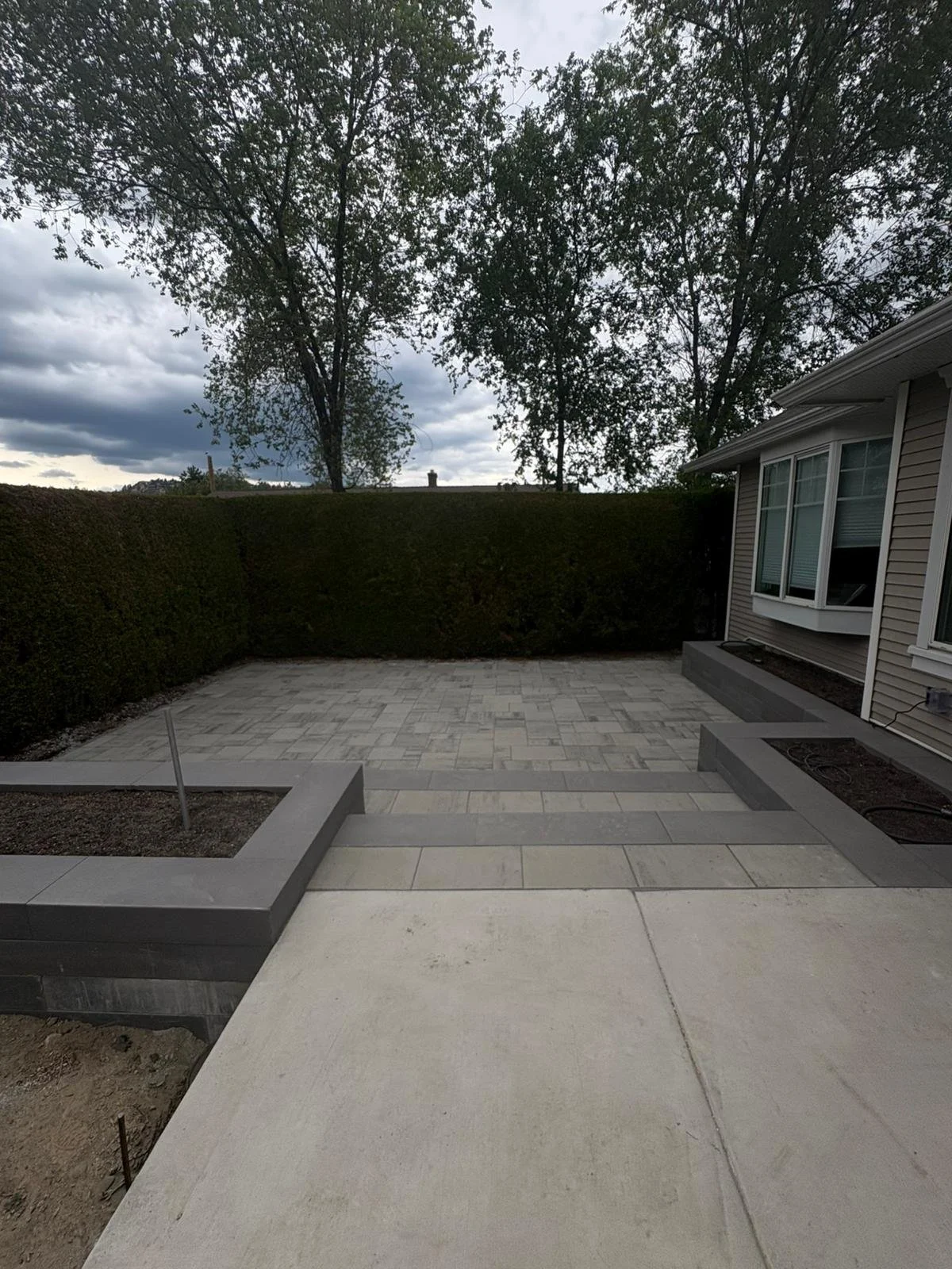 Newly paved backyard patio with concrete and pavers, bordered by a house with beige siding and large windows, surrounded by a tall hedge and trees under a cloudy sky.