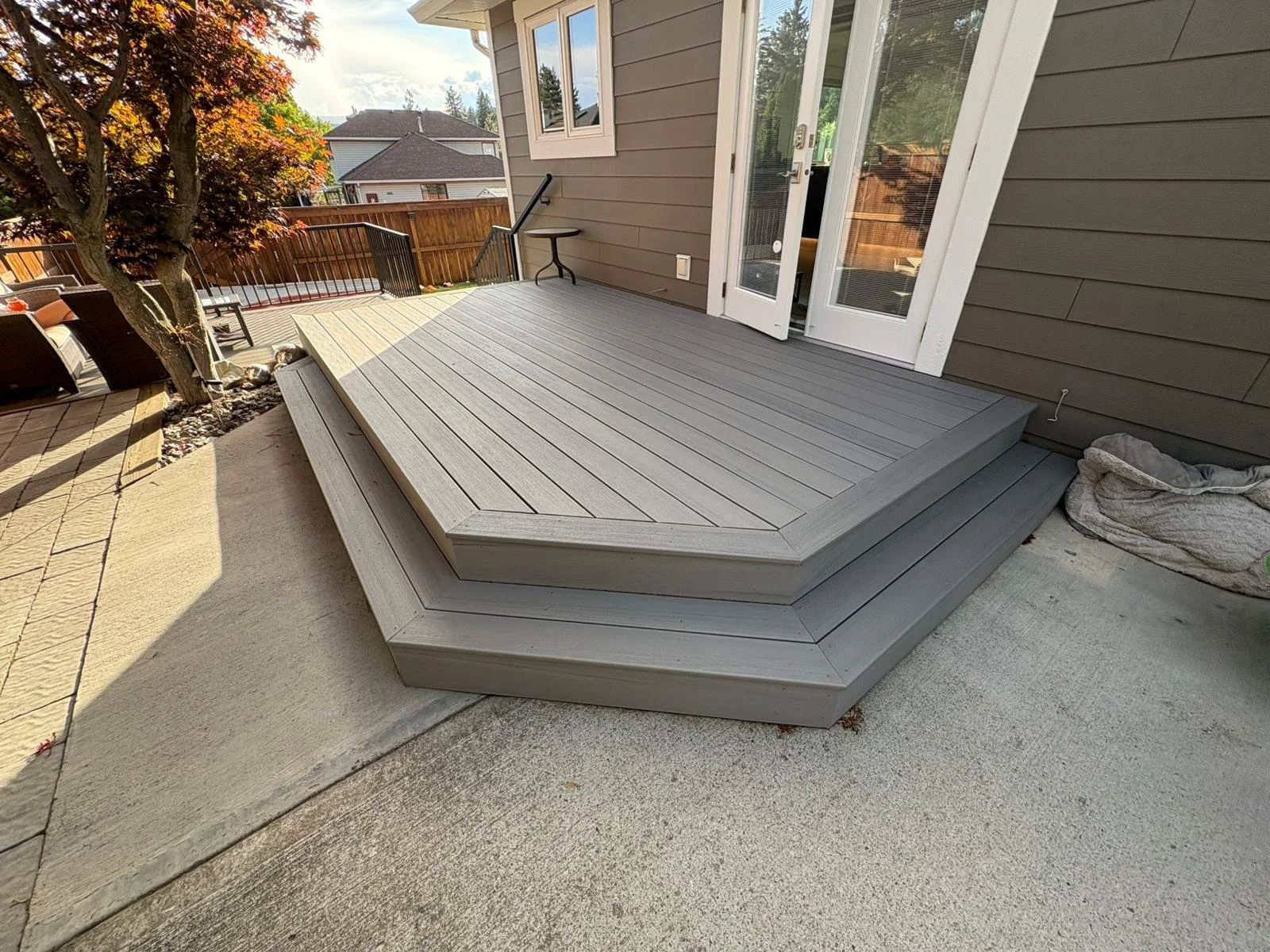 Gray wooden deck attached to a house with a glass door. Surrounding elements include a tree, outdoor furniture, a retaining wall, and neighboring houses.